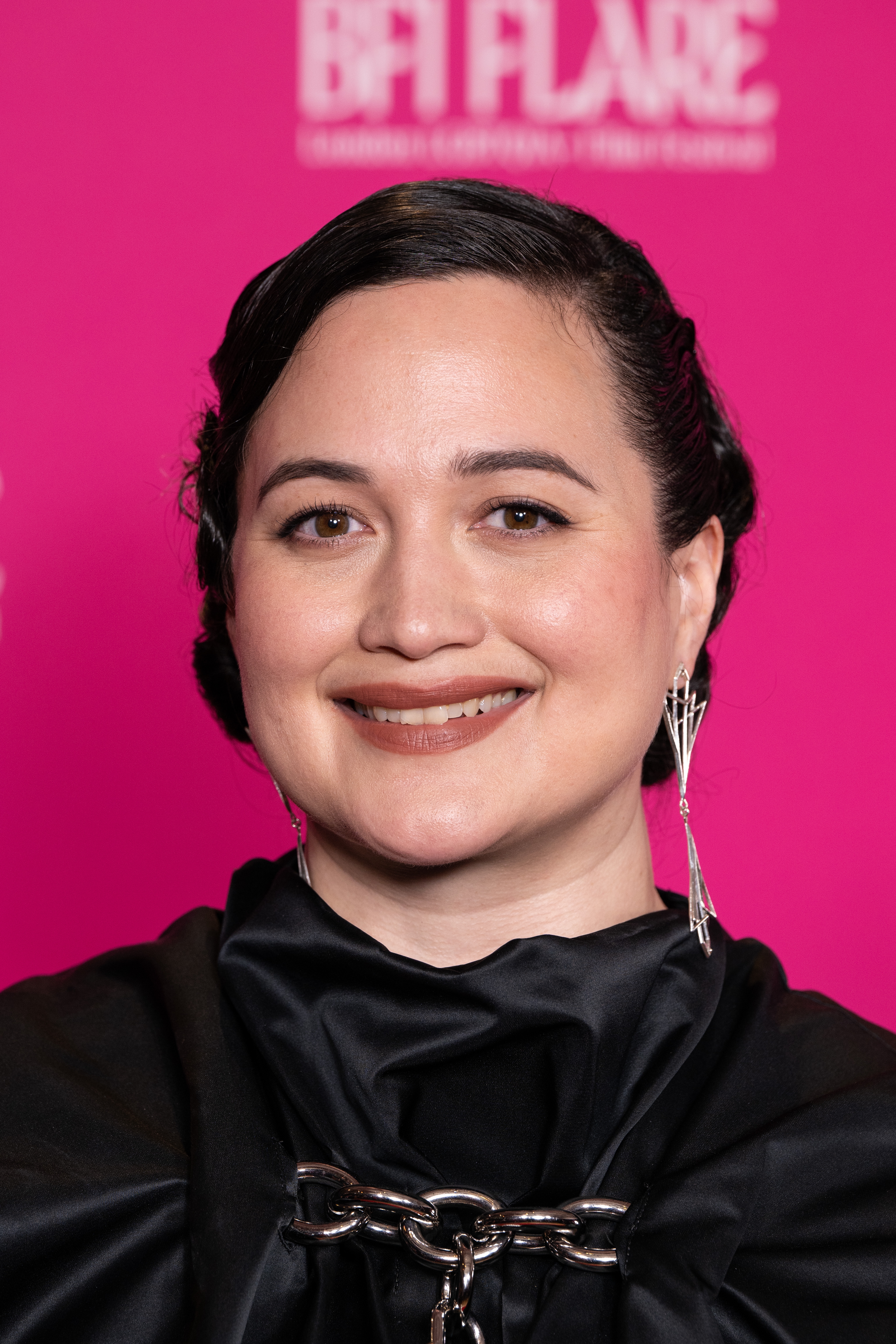 Lily Gladstone smiling, wearing a black top with metal chain detail and geometric earrings. Background has BFI Flare text