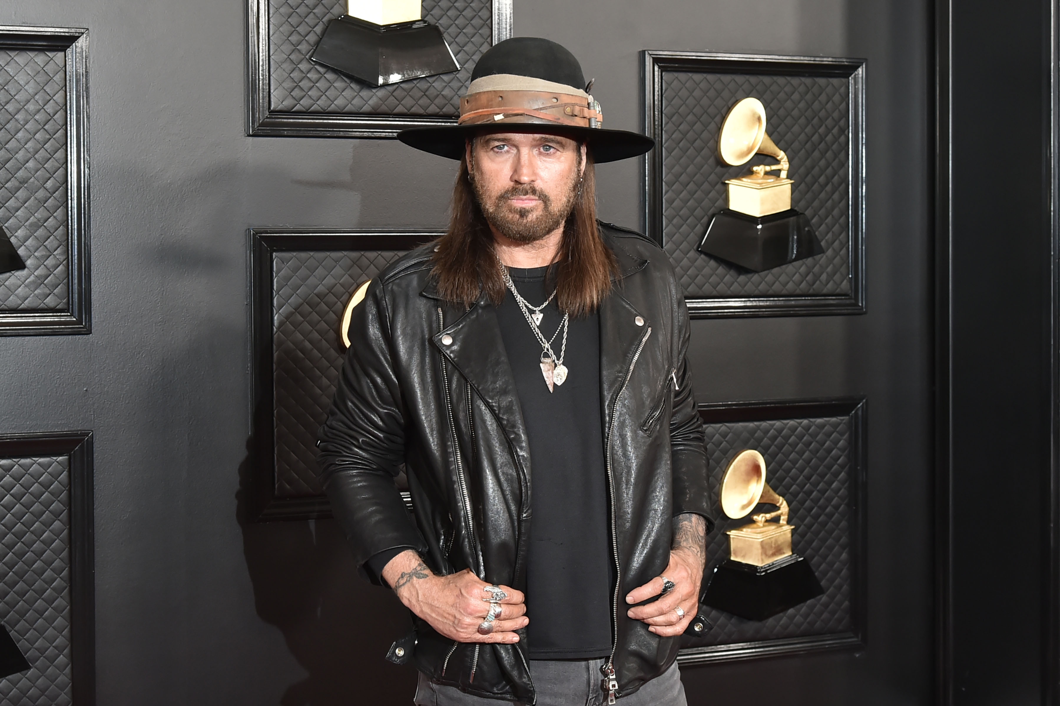 Billy Ray Cyrus in a leather jacket and wide-brim hat poses on a music awards red carpet, with Grammy statues displayed on the wall behind