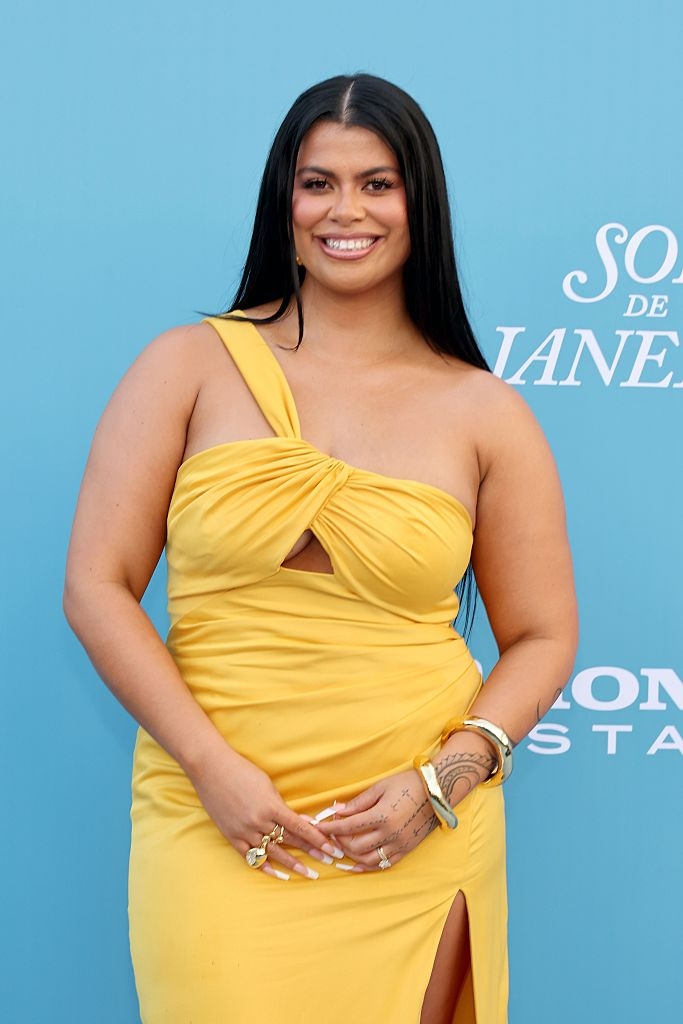 Person in a one-shoulder gown with subtle jewelry, smiling at a formal event. Blue background with partial text "Sons de Janeiro."