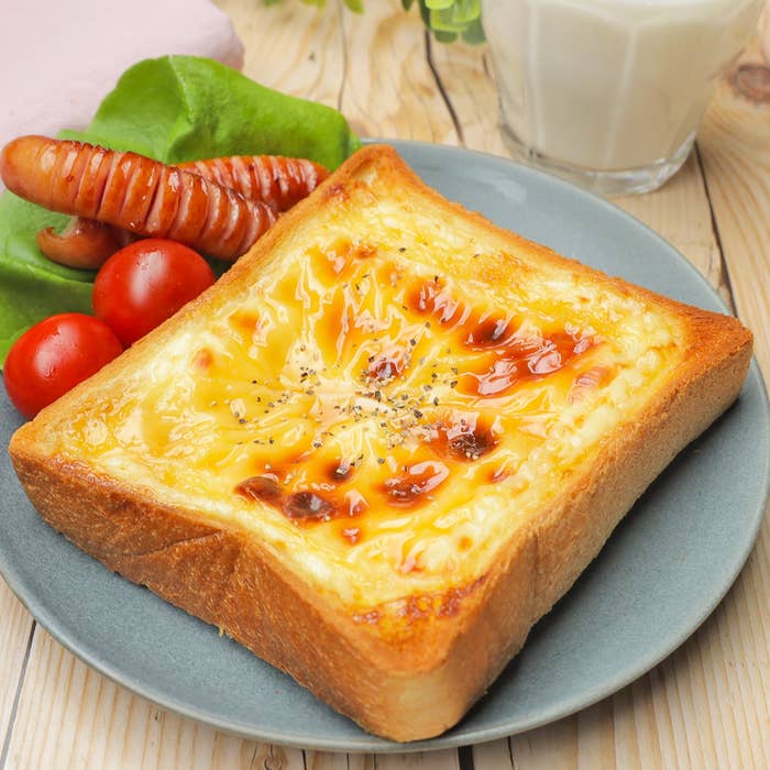 トーストにとろけたチーズとソース、付け合わせにソーセージとトマト、奥にグラスの牛乳がある朝食シーン。