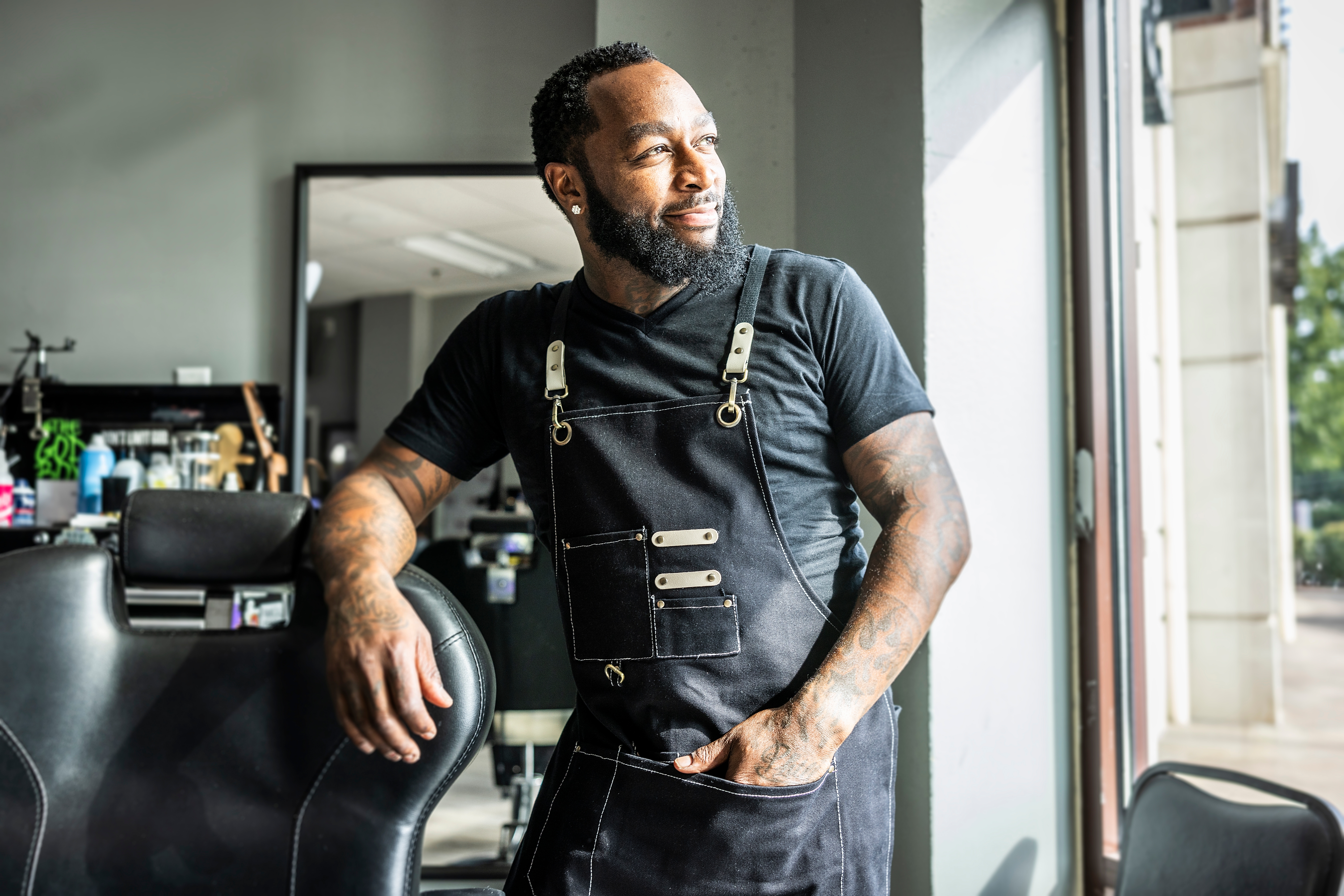 A person in a barbershop wearing a black apron over a t-shirt, looking thoughtfully out the window with one hand in their apron pocket