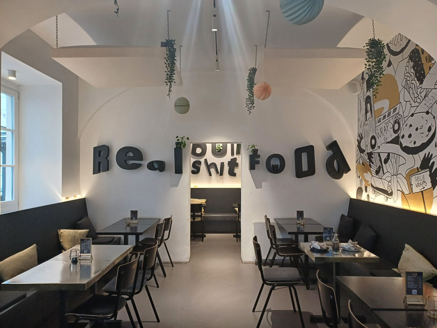 Cafe interior with minimalist decor; walls feature the words "Real Shit Food" due to overlapping letters on both walls. Tables and chairs are neatly arranged