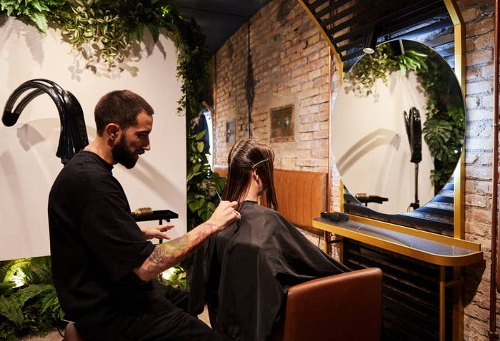 A hairstylist cuts a client's hair in a chic, plant-filled salon with a brick wall and large round mirror