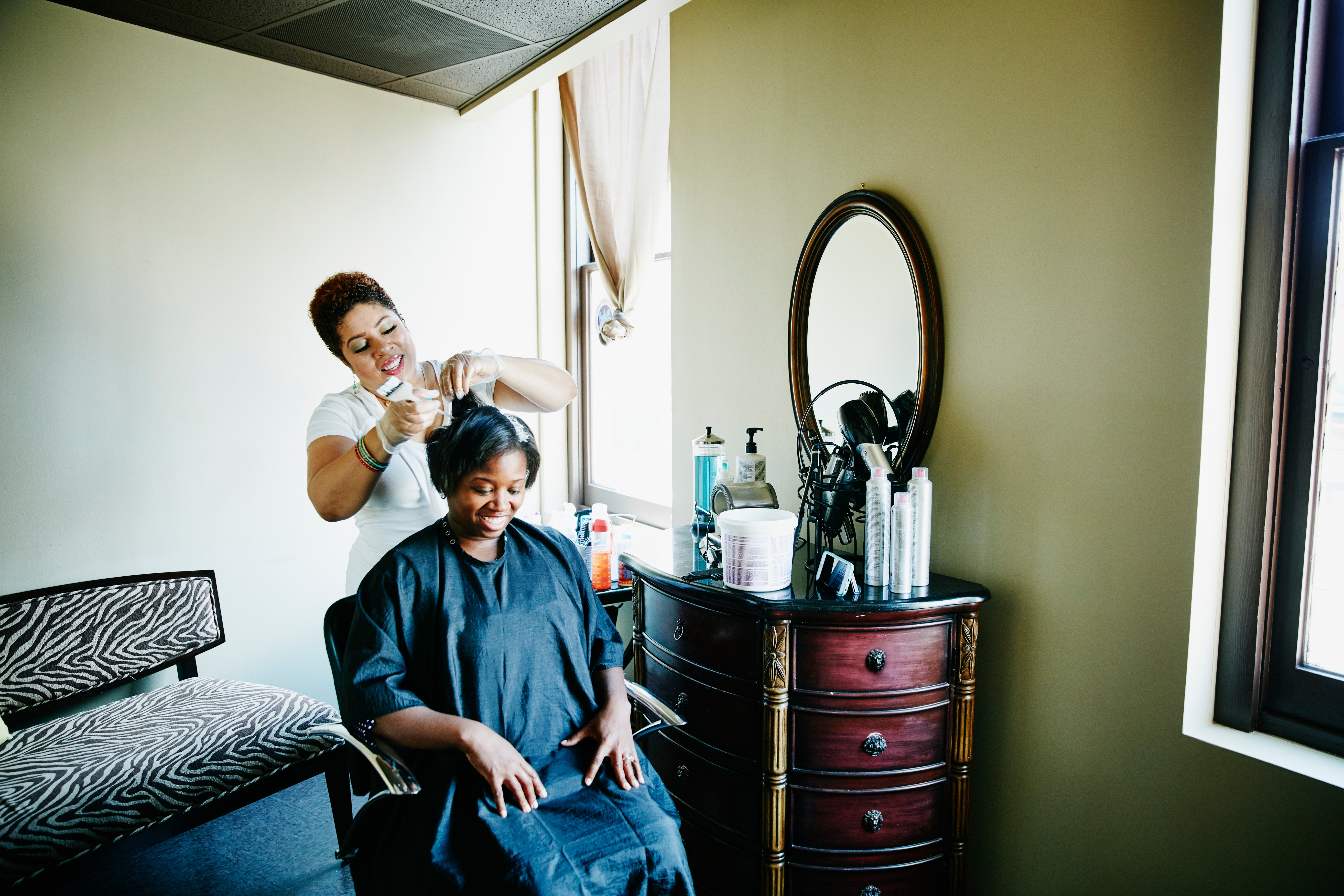 A hairstylist joyfully trims a seated client's hair in a cozy, sunlit salon with a zebra-print chair and an oval mirror