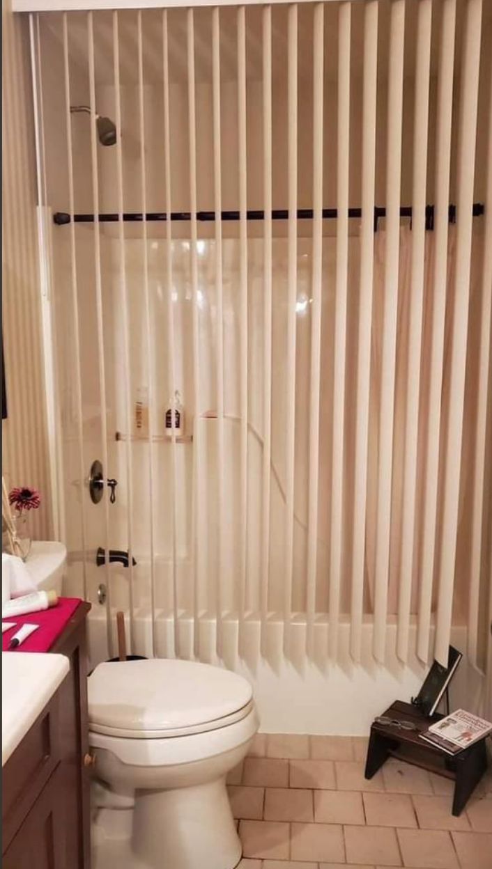 Bathroom with vertical blinds instead of a shower curtain over the bathtub, next to a toilet and a small table with a magazine