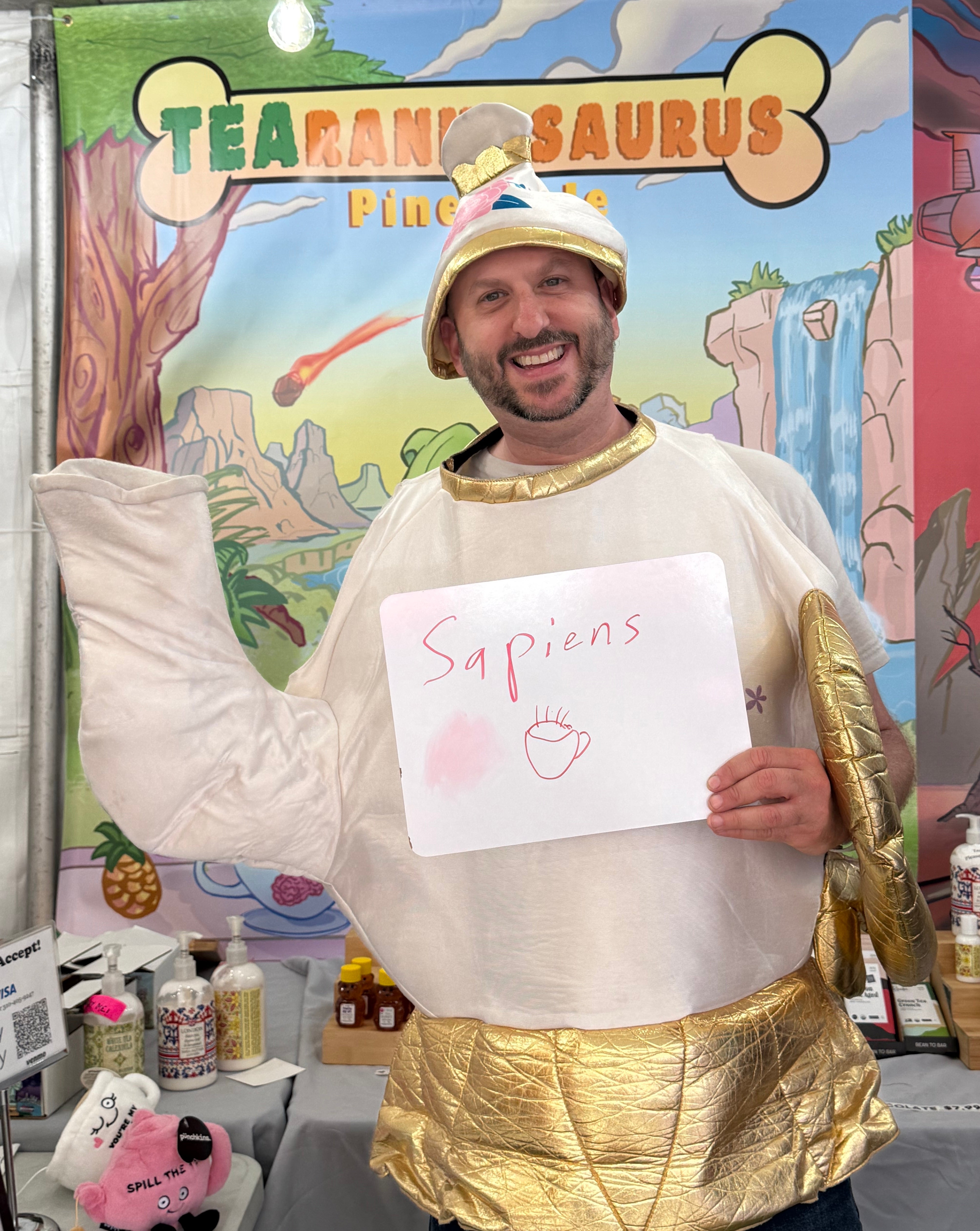 Person in a teapot costume holds a sign with "Sapiens" and a hand drawing, standing in front of a stall with various drink products