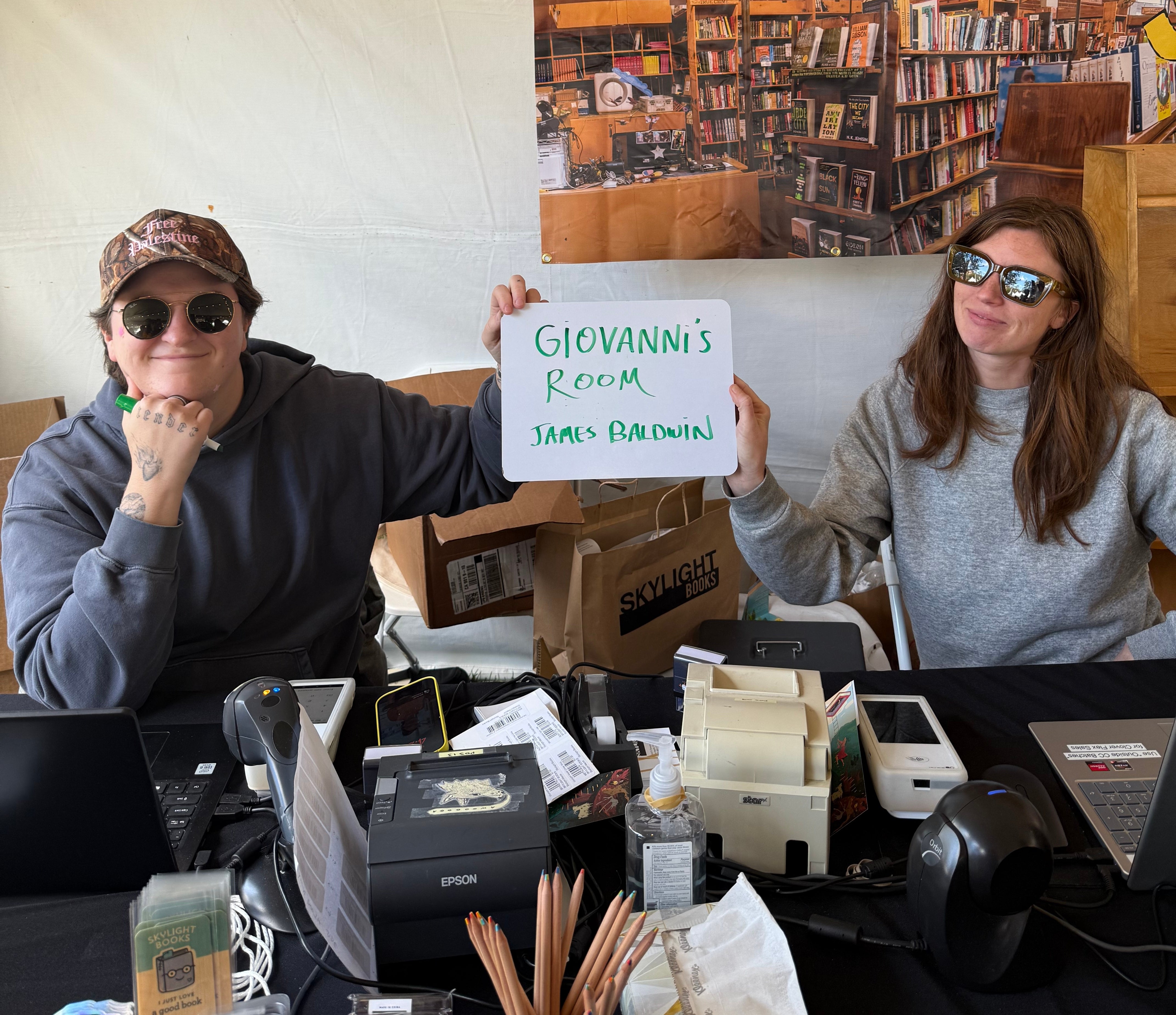 Two people sit at a table in a bookstore. One holds a sign that says "Giovanni's Room, James Baldwin."