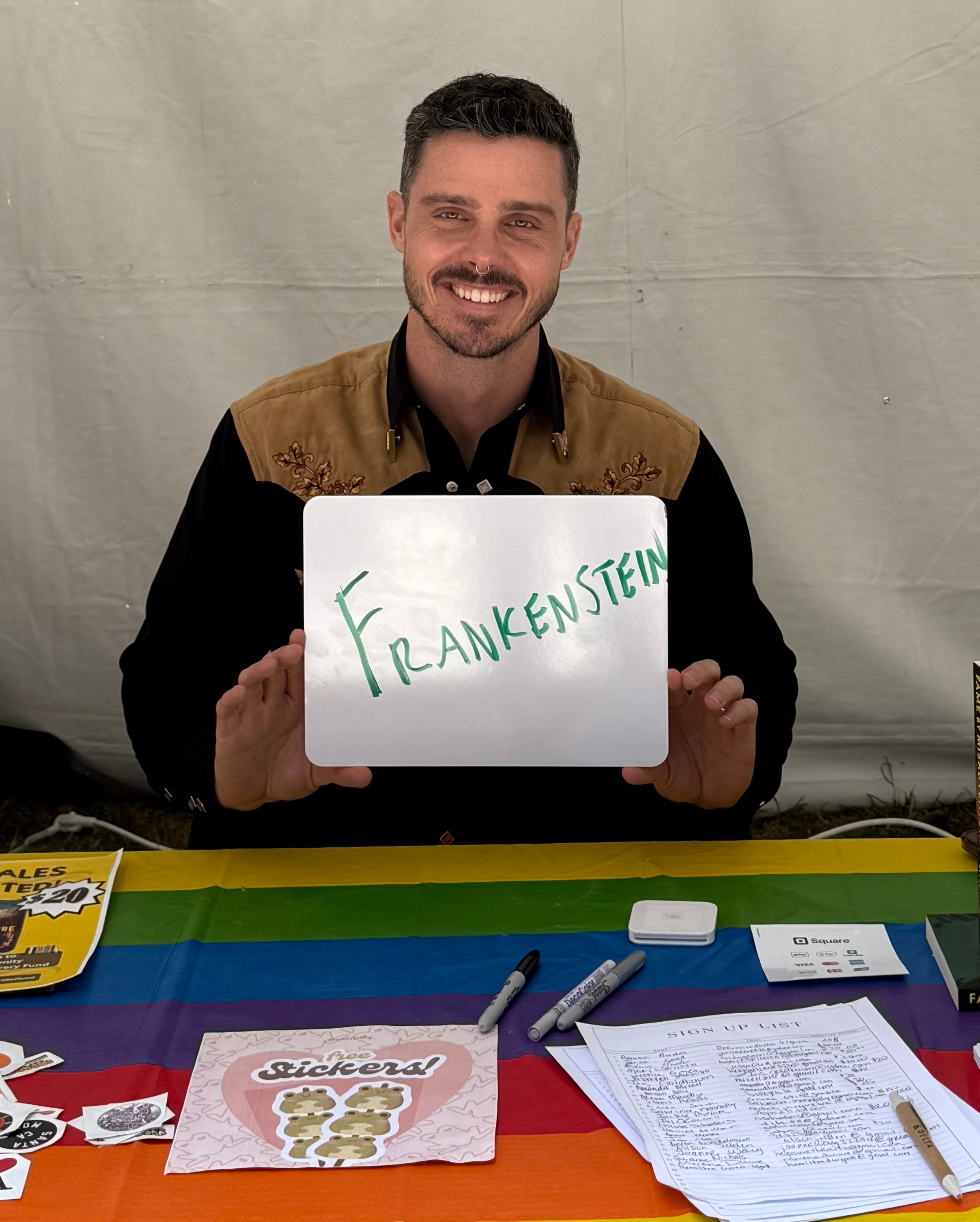 Person smiling, holding a whiteboard with "Frankenstein" written on it, sitting at a table with stickers and pens