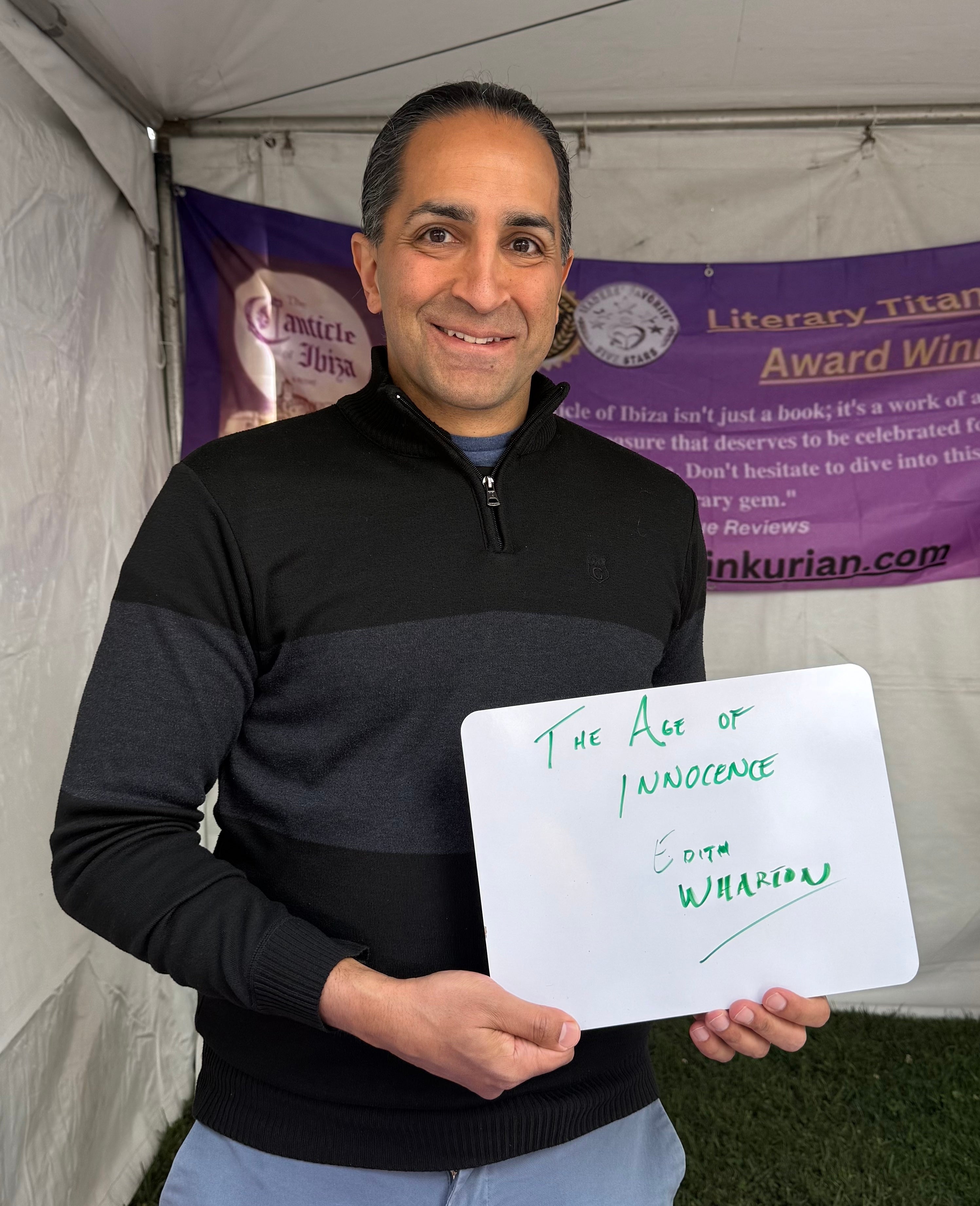 Person holding a whiteboard with "The Age of Innocence, Edith Wharton" written on it. Banner and tent visible in background