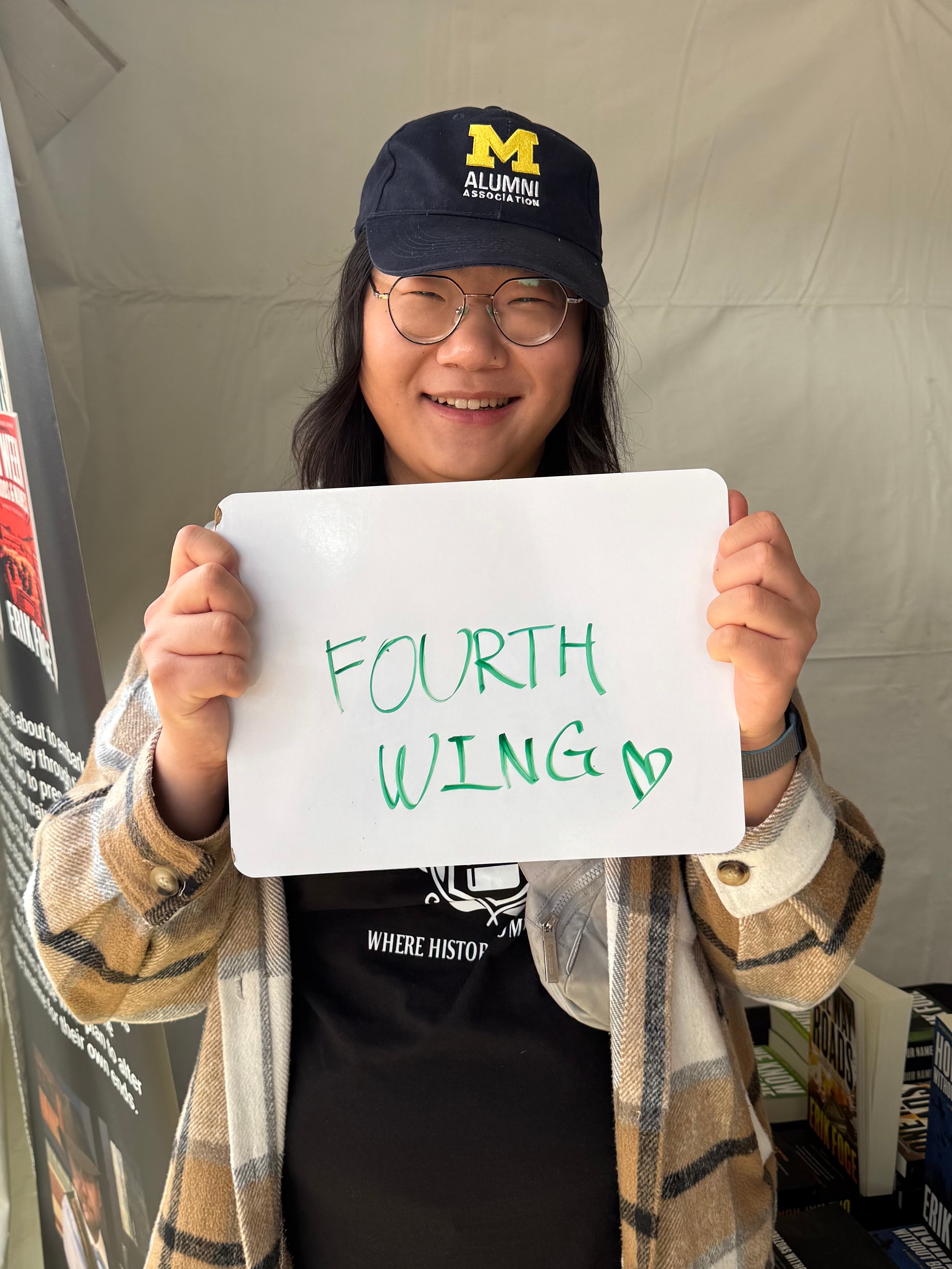 Person holding a whiteboard with "FOURTH WING" written on it, wearing a checkered jacket and a cap that reads "M ALUMNI."