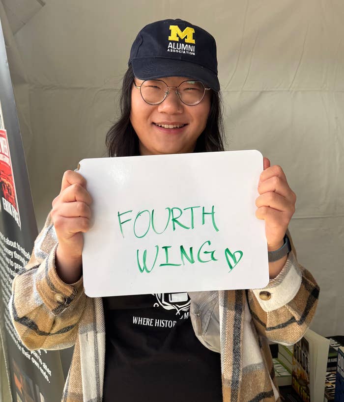 Person holding a whiteboard with "FOURTH WING" written on it, wearing a checkered jacket and a cap that reads "M ALUMNI."