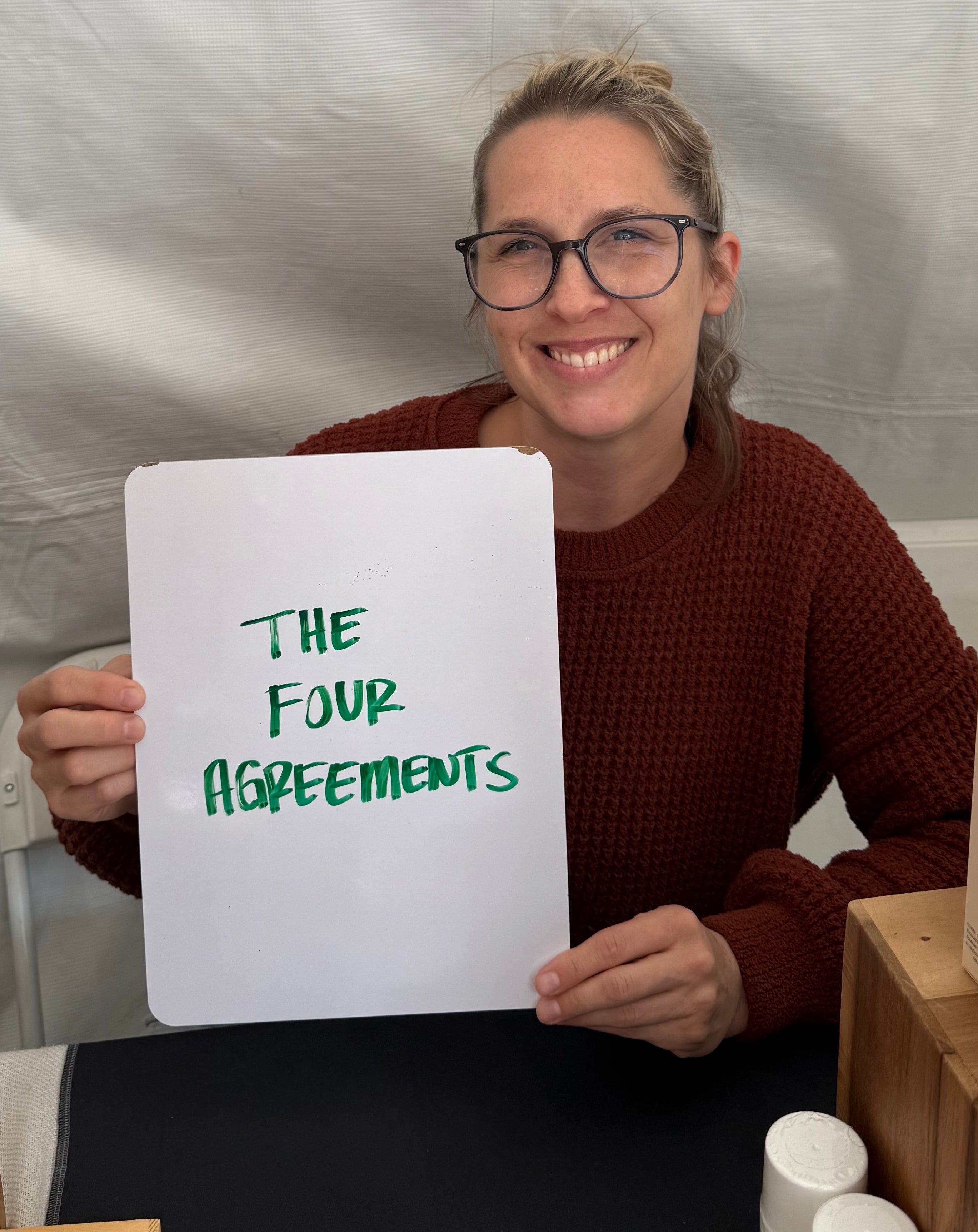 Person smiling, holding a sign that reads "The Four Agreements," with boxes and bottles on the table in front of them