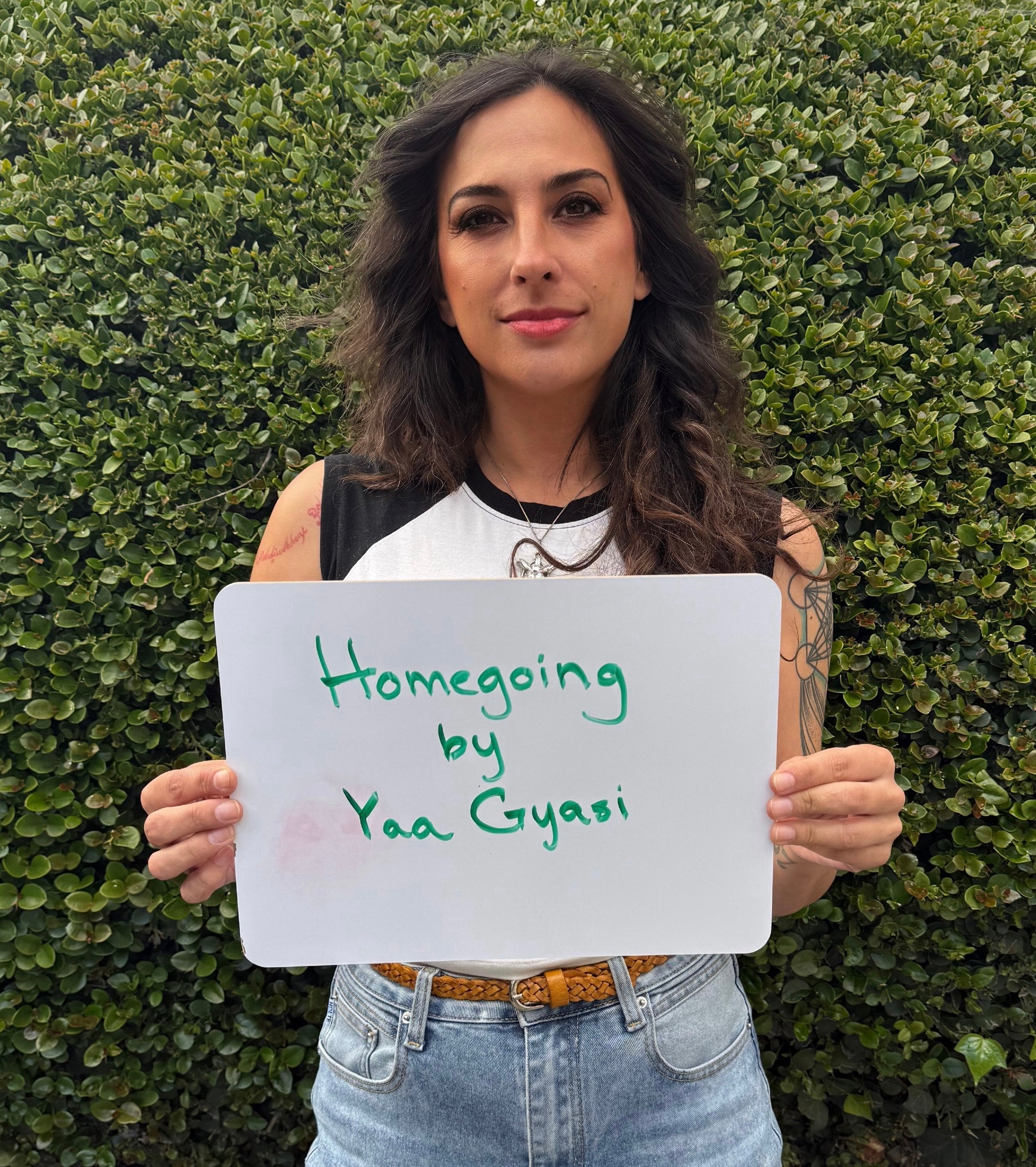 Person holding a sign with the book title "Homegoing" by Yaa Gyasi, standing in front of greenery