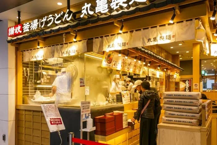 うどん店の外観。カウンターでお客さんが注文し、スタッフが調理中。看板には店名が表示されています。