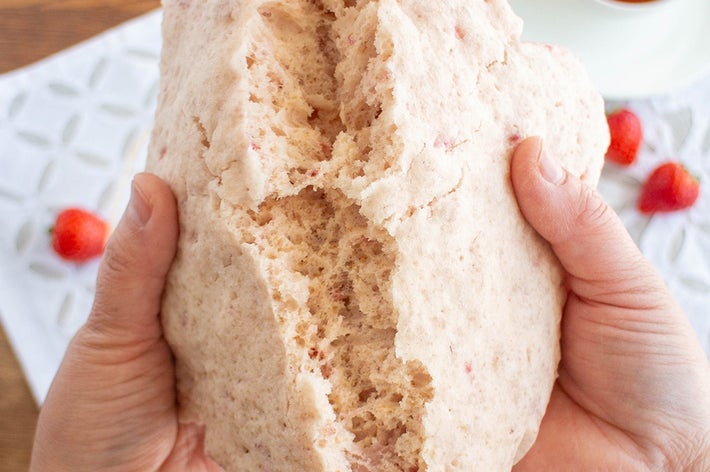 手で割られたスコーンの断面が見えている。背景には紅茶のカップとラズベリーがある。