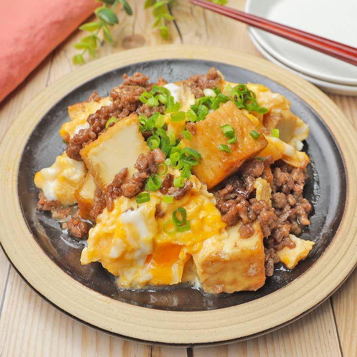 焼き豆腐とひき肉、卵が絡む料理、ネギがトッピングされています。白い皿に盛られ、周囲には箸と食器があります。