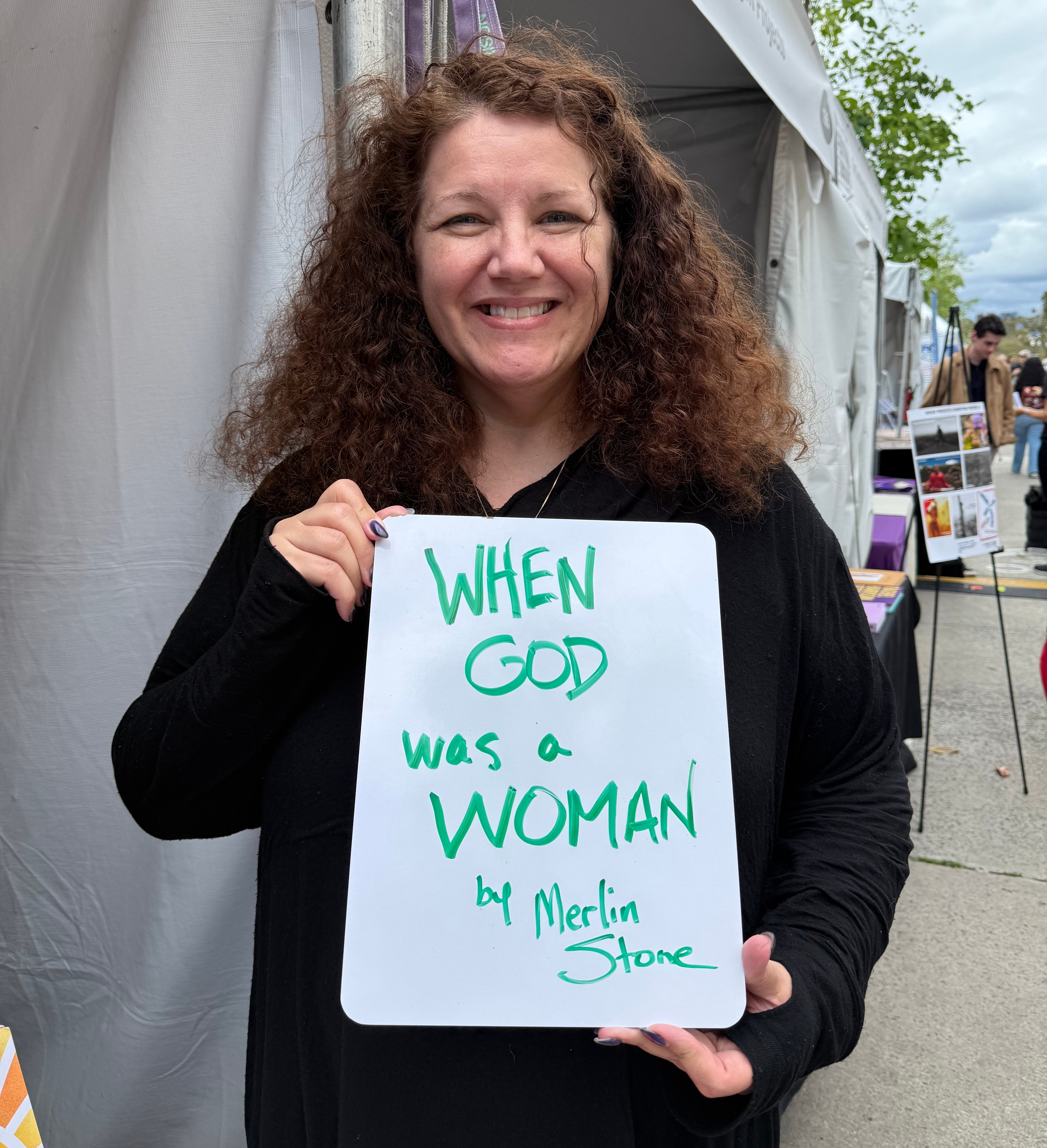 Person smiling and holding a sign that reads "When God was a Woman by Merlin Stone."