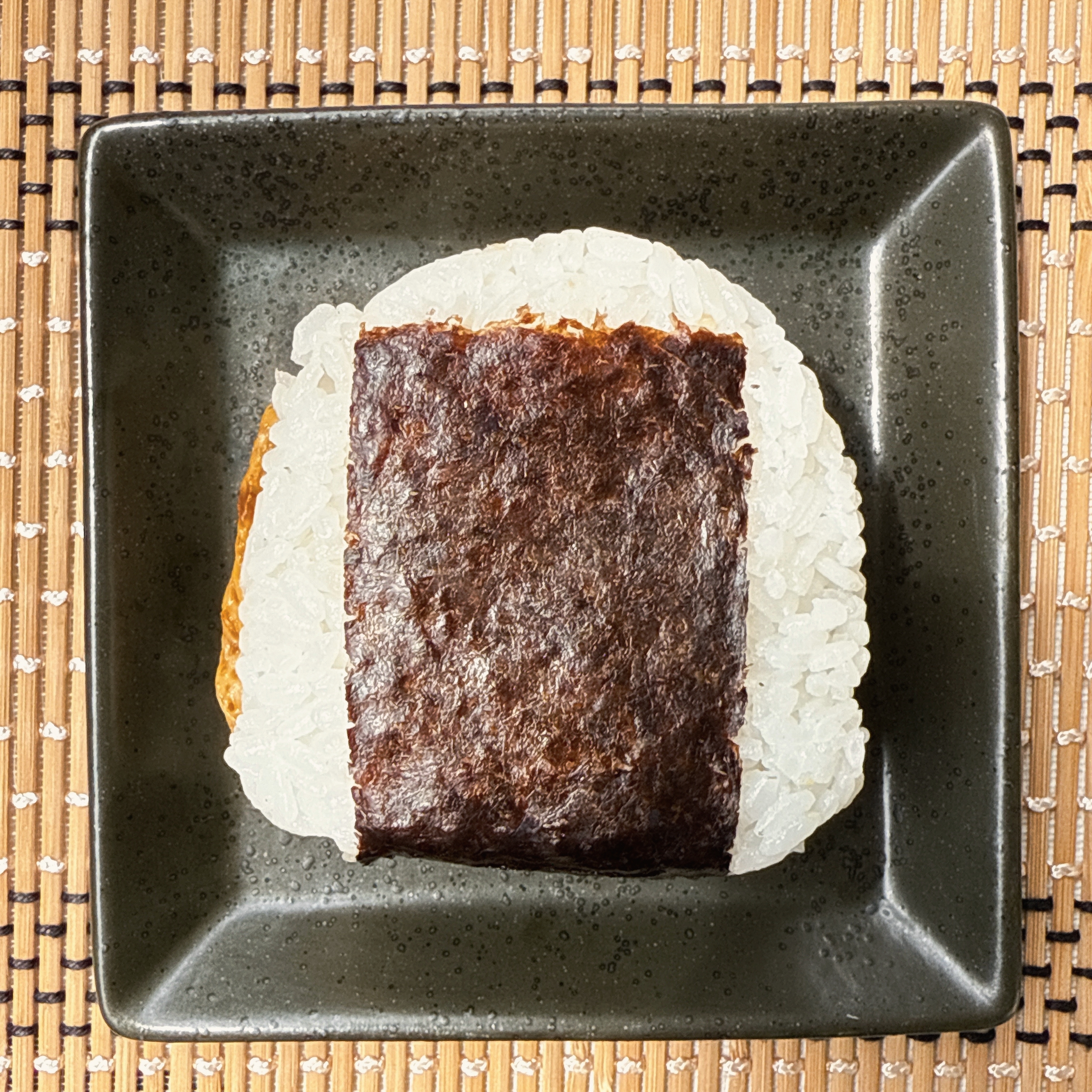 海苔で包んだおにぎりが黒い皿に載せられ、編まれたマットの上に置かれています。