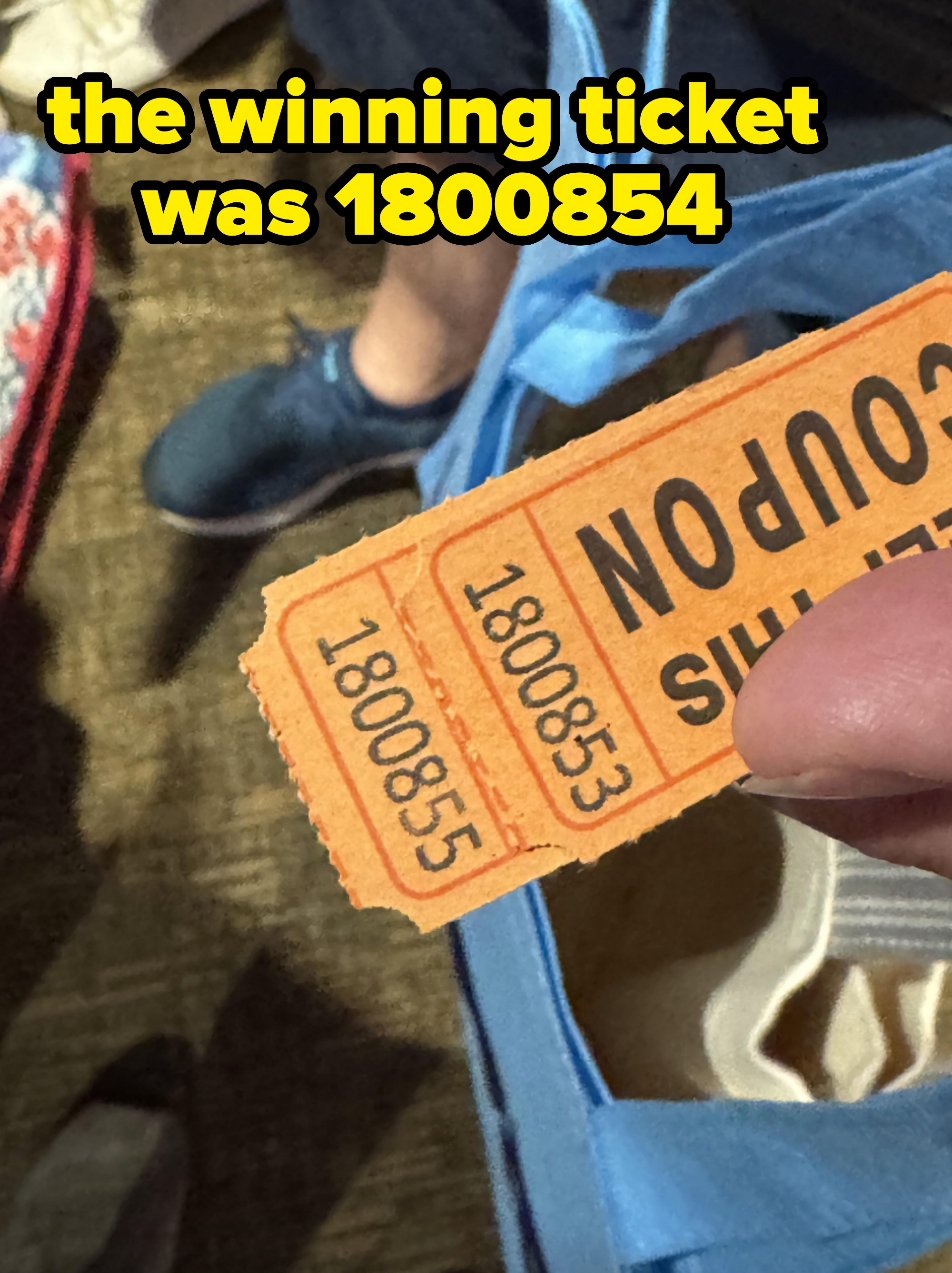 Person holding orange raffle ticket, standing among others with visible legs. Blue bag partially seen