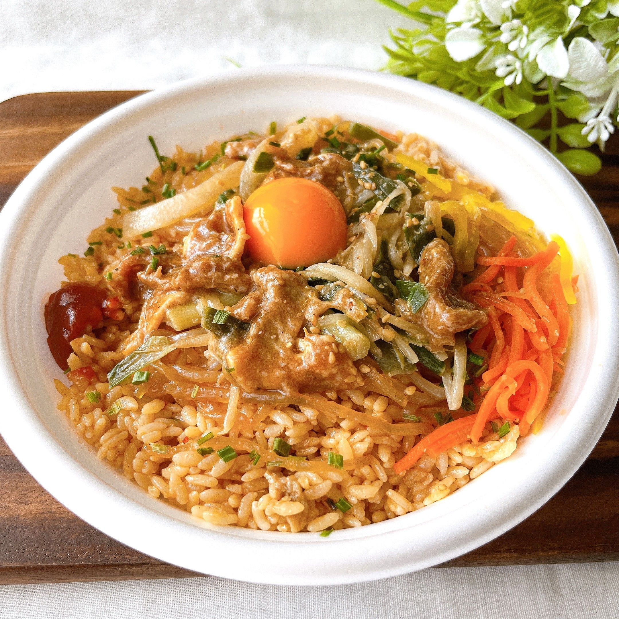 ボウルに盛られたビビンバ。焼肉、卵黄、野菜がご飯に乗っている。白い皿と背後に花が見える。