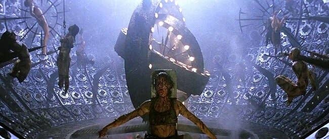 A person covered in dark liquid stands in a futuristic room with suspended figures around and a large, rotating mechanism above