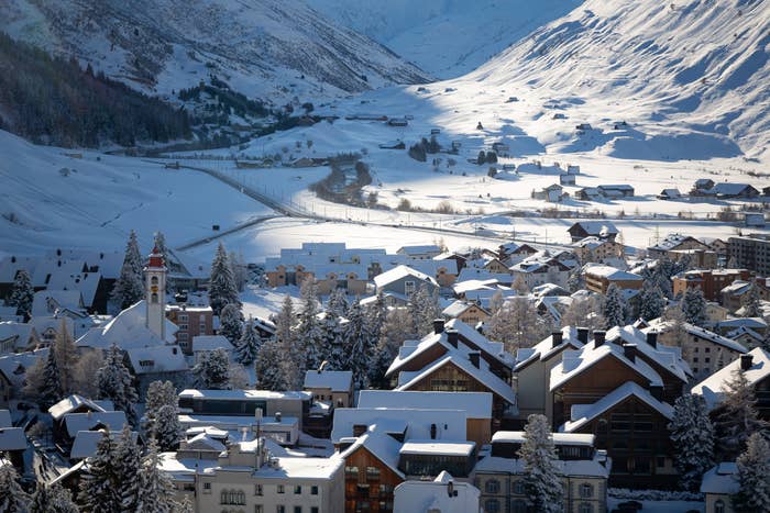 Snow-covered village with mountain backdrop, showing rooftops, a church tower, and trees, creating a serene winter scene