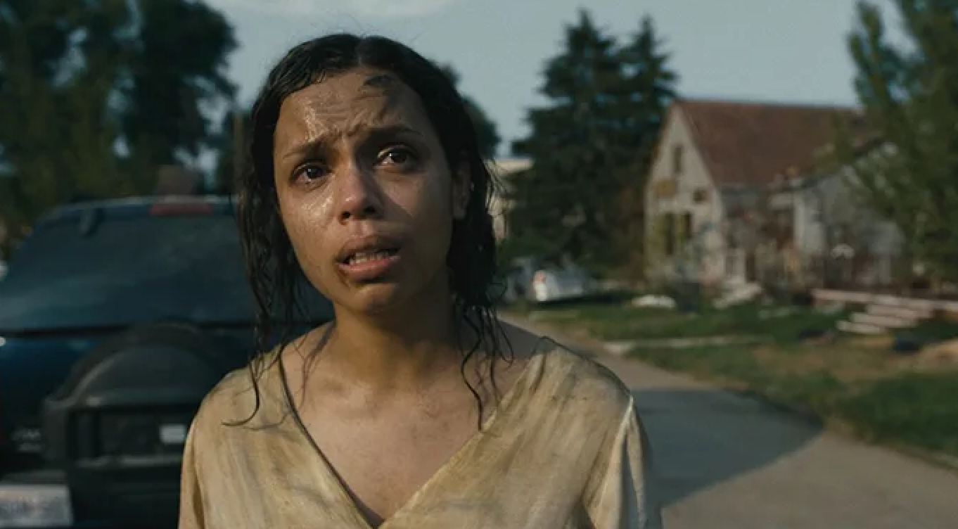 Person outdoors with a distressed expression, wearing a worn-out shirt. Houses and a parked car are visible in the background
