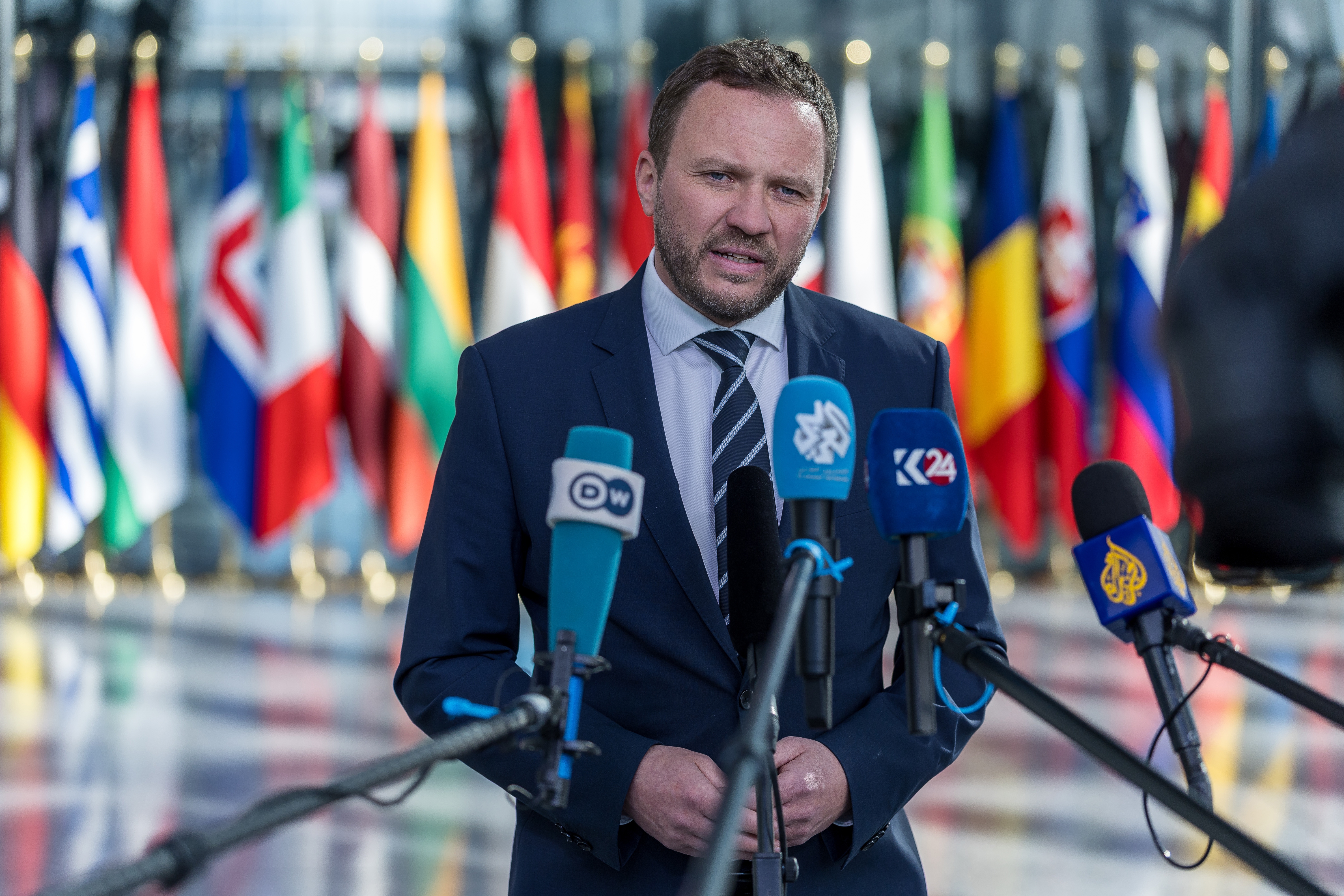 A man in a suit giving a press conference, standing in front of multiple microphones with various international flags in the background