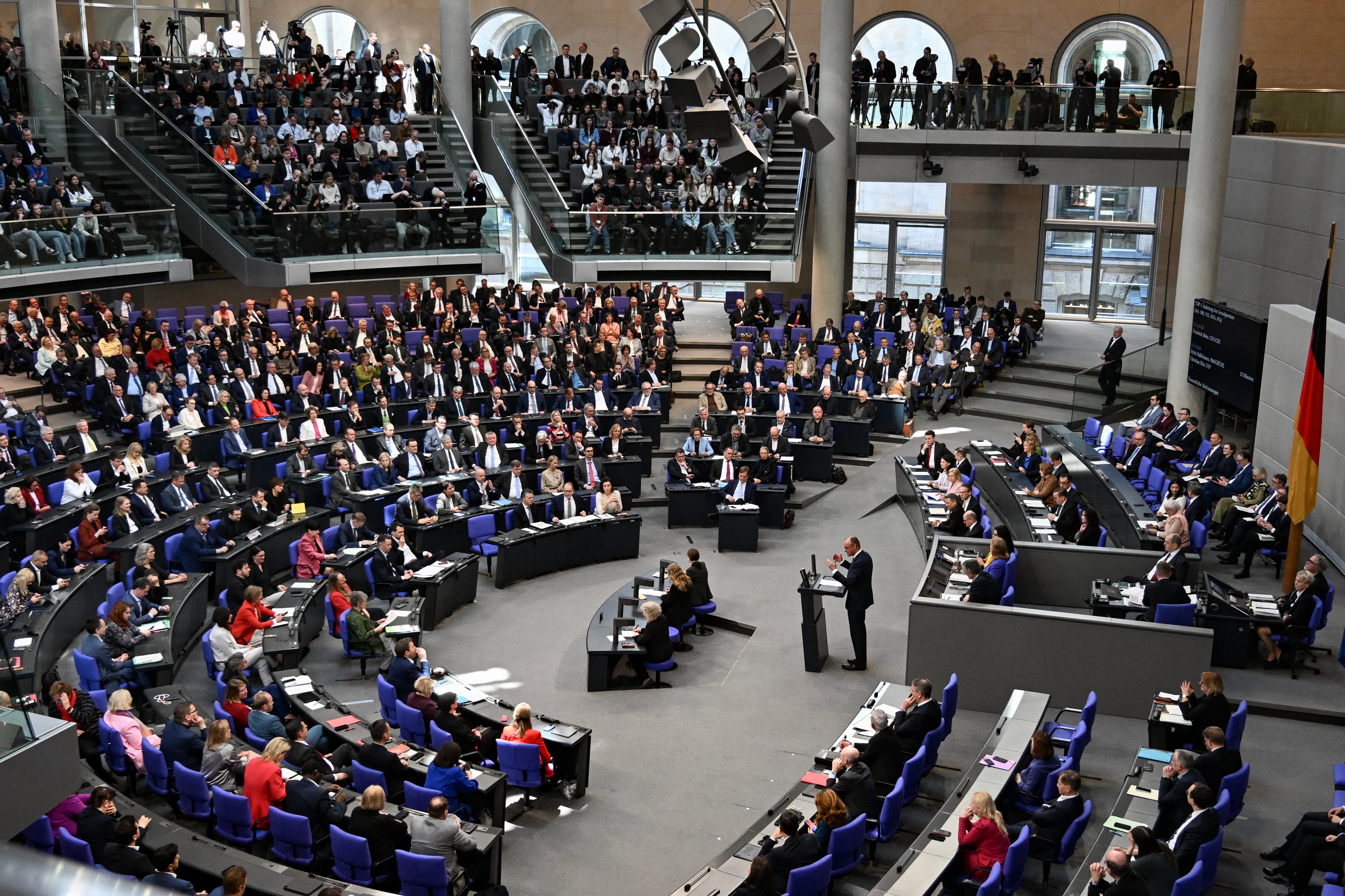 Large assembly meeting in a modern hall, attendees seated in a semi-circle while a person speaks at a podium, viewed from above