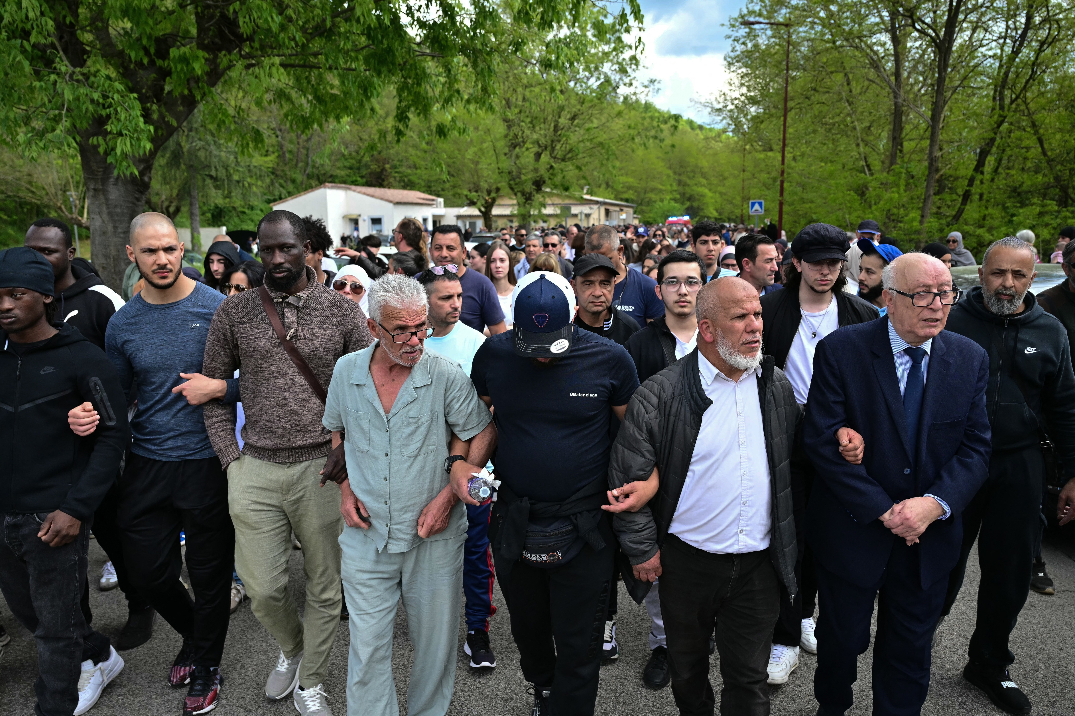 A diverse group of people linked arm in arm, marching in solidarity down a tree-lined street