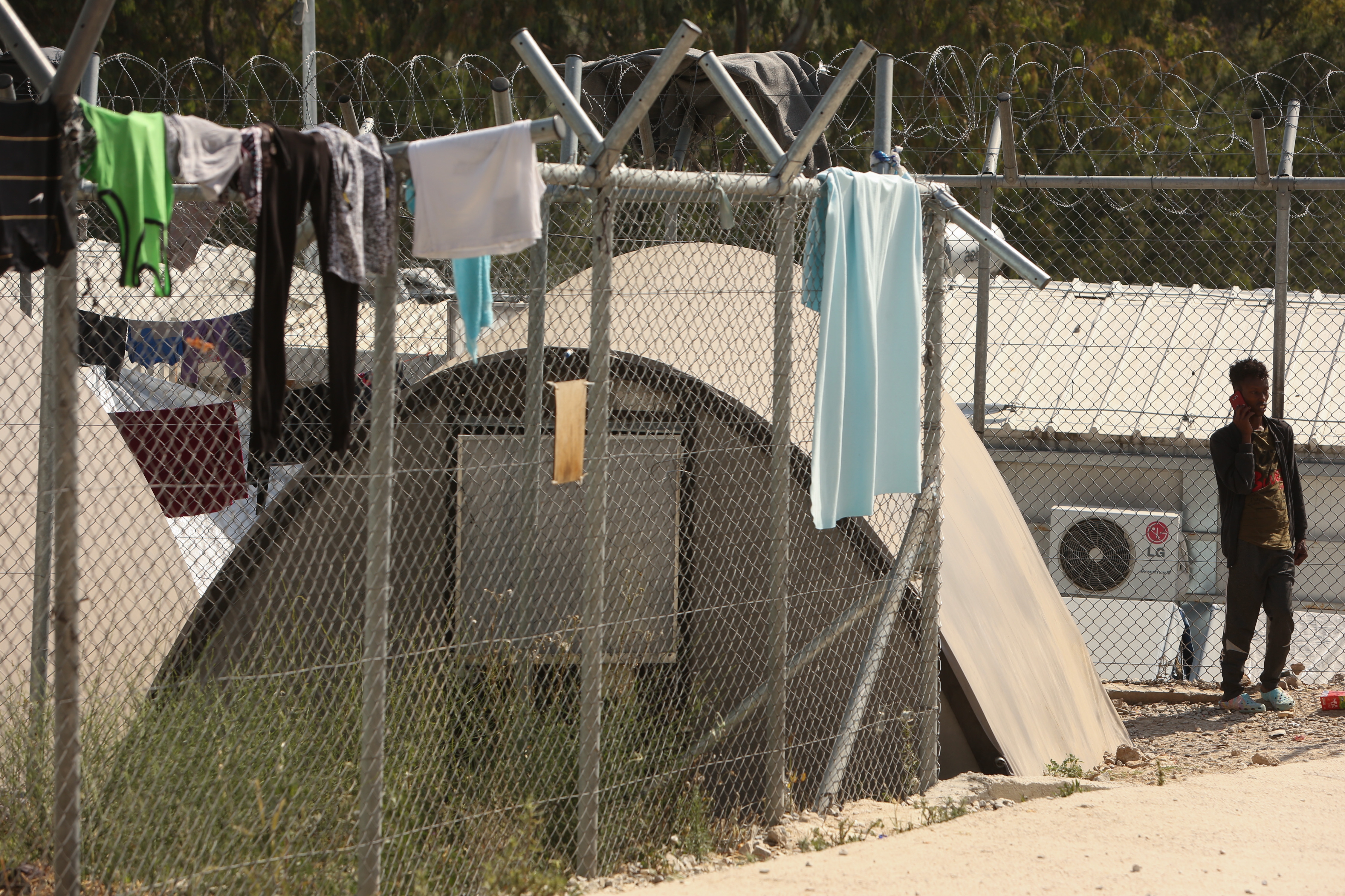 Clothing hangs on a fence at a refugee camp with tents and barbed wire. A person stands nearby, looking at the scene