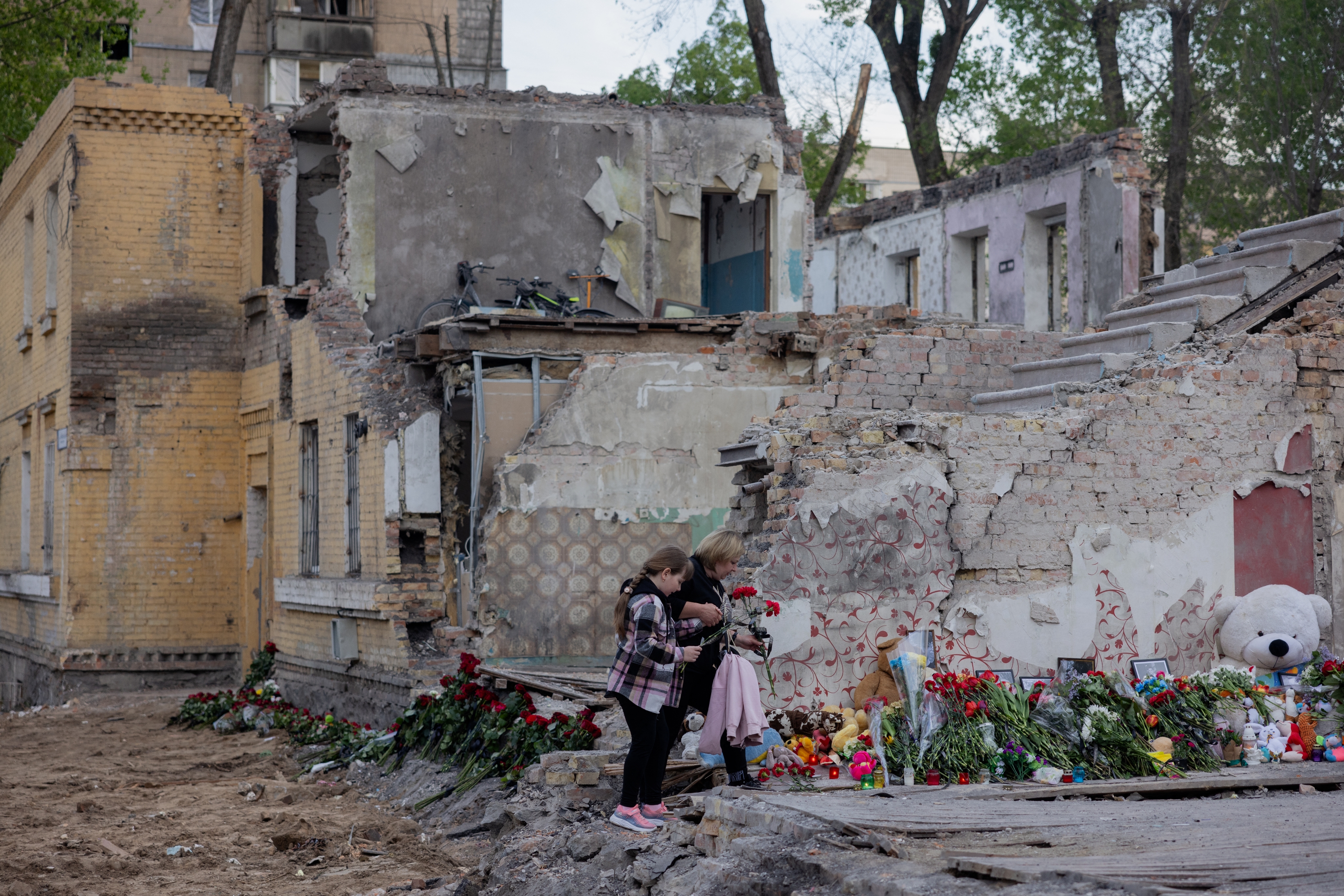Two people stand by a memorial with flowers and plush toys in front of a damaged building