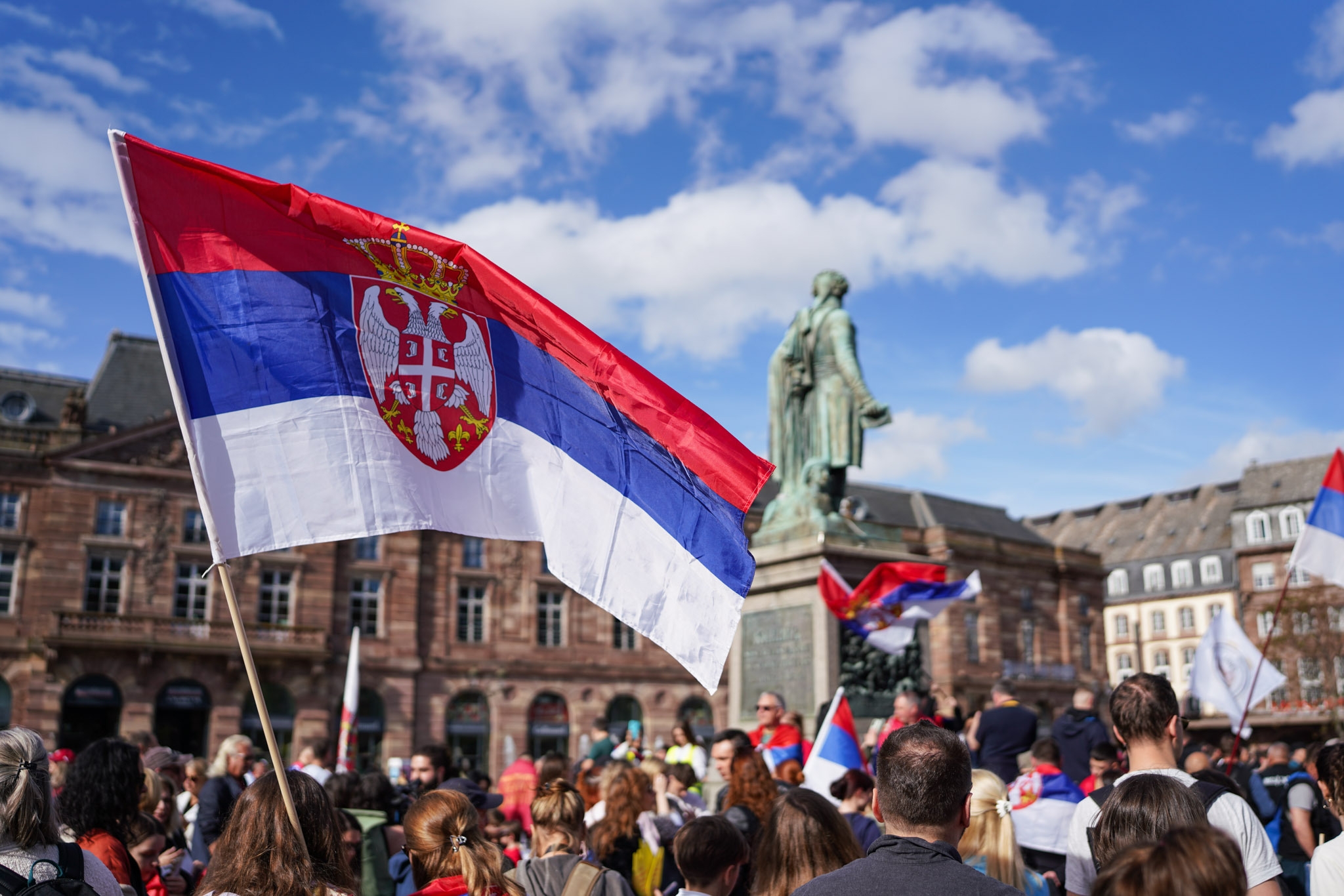People gather in a plaza holding Serbian flags, near a statue, during a public event or protest