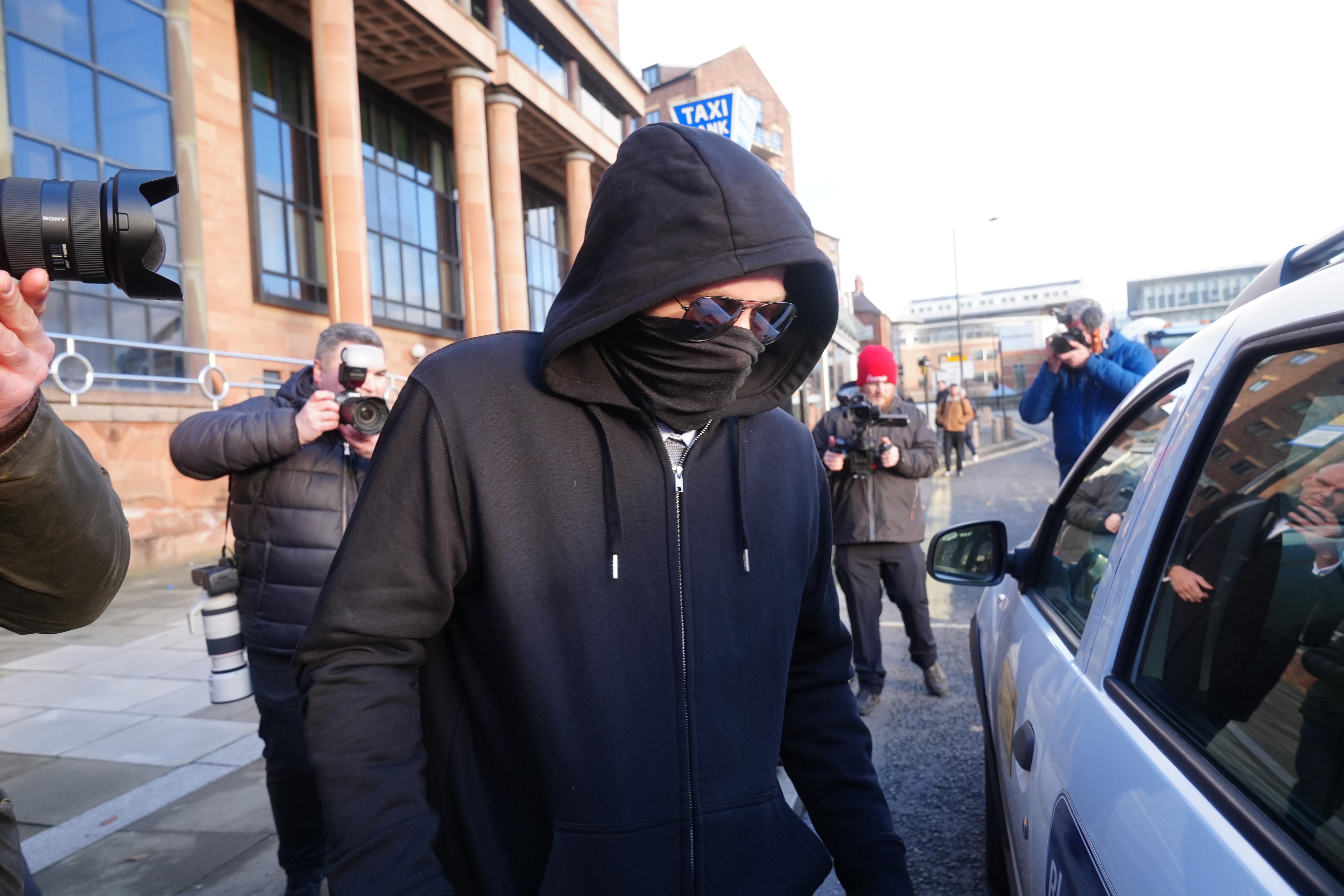 Person in a hoodie and face covering walks past photographers outside a building