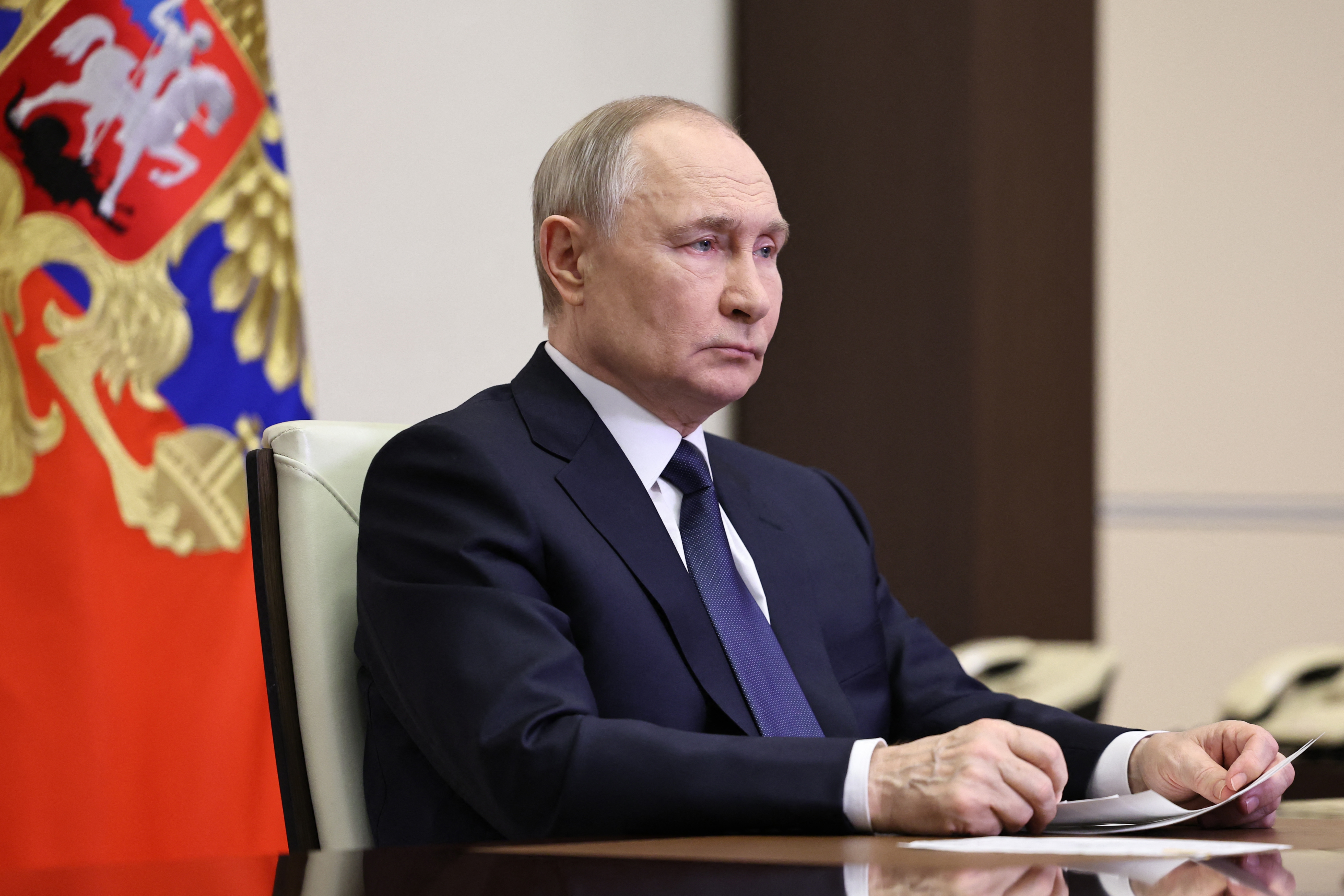 Vladamir Putin sits at a desk with a document, a flag with an emblem in the background