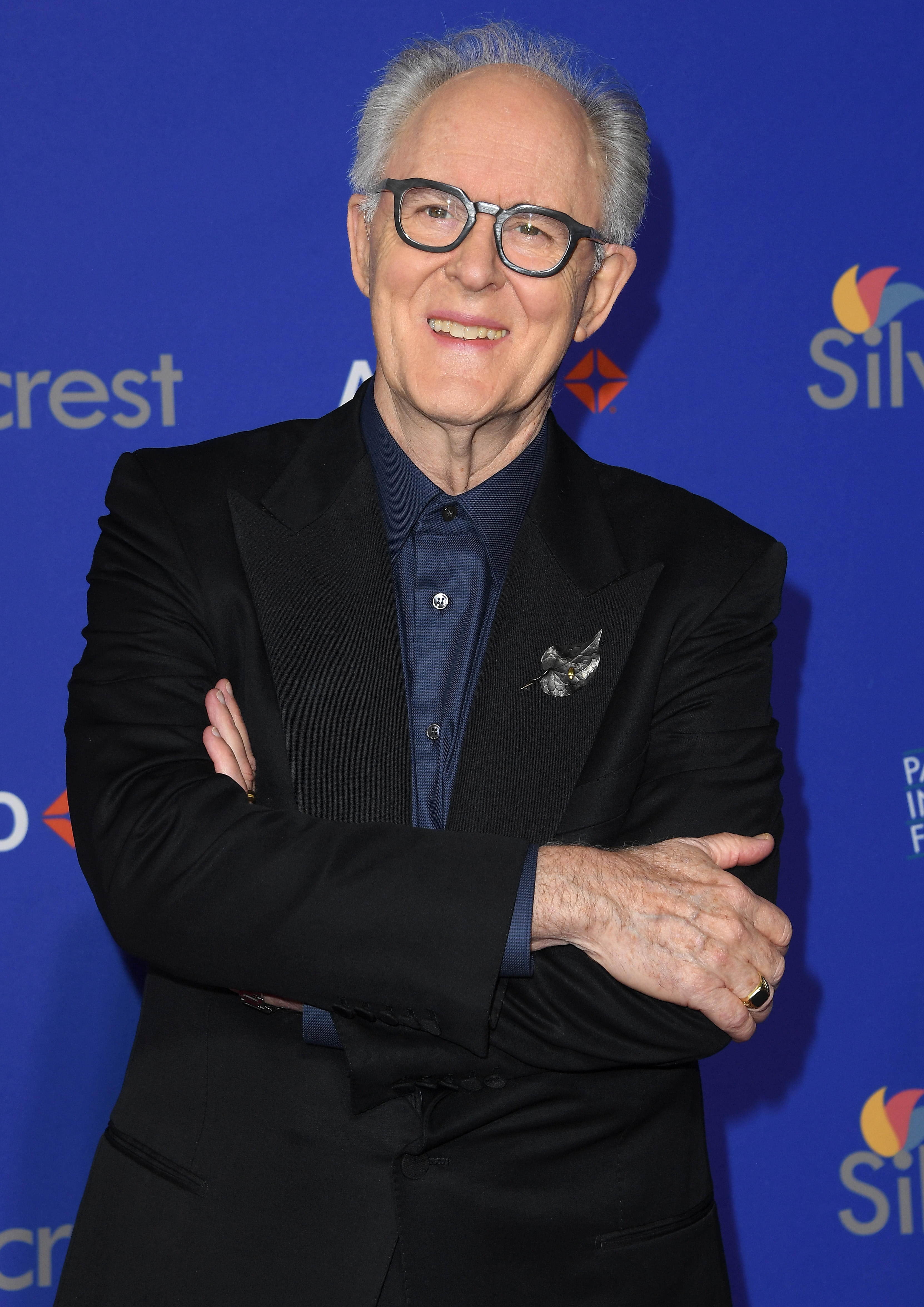 John Lithgow in a dark suit with a bird pin, smiling and posing with arms crossed on a blue background at a formal event