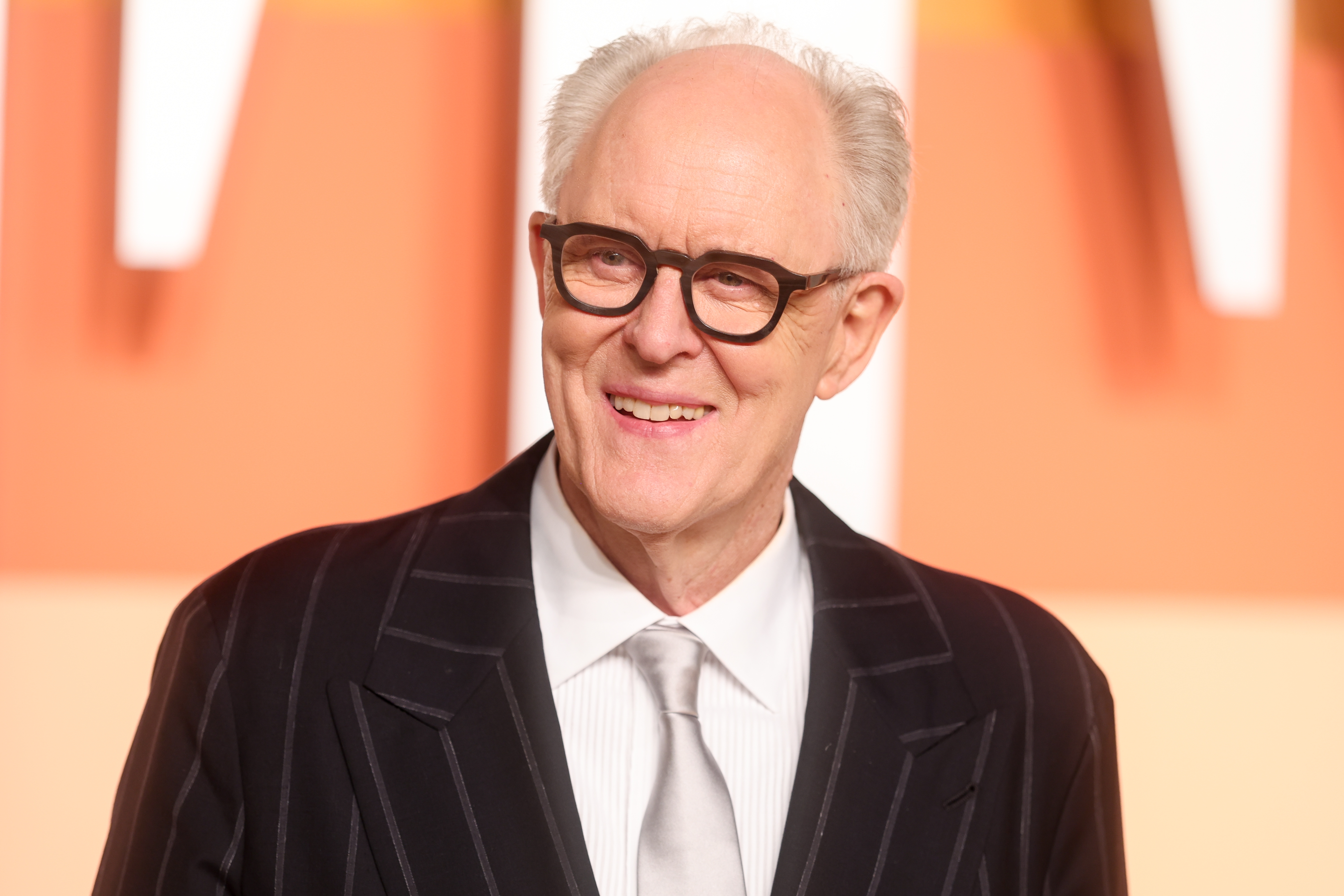 John Lithgow in a pinstriped suit and tie is smiling on a formal event backdrop