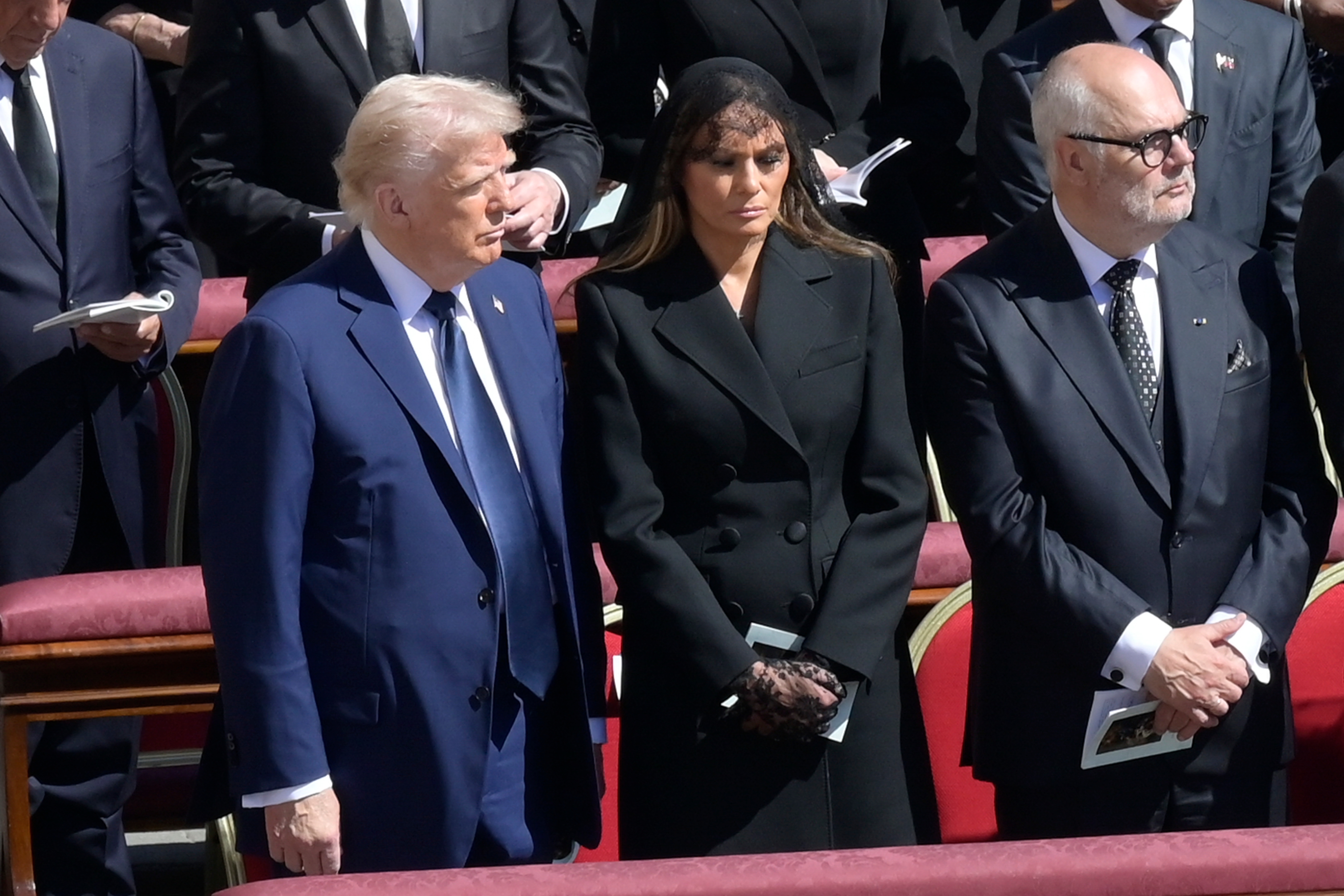 Three people stand at a formal event, with one wearing a dark suit, another in a black coat and veil, and the third in a suit with glasses