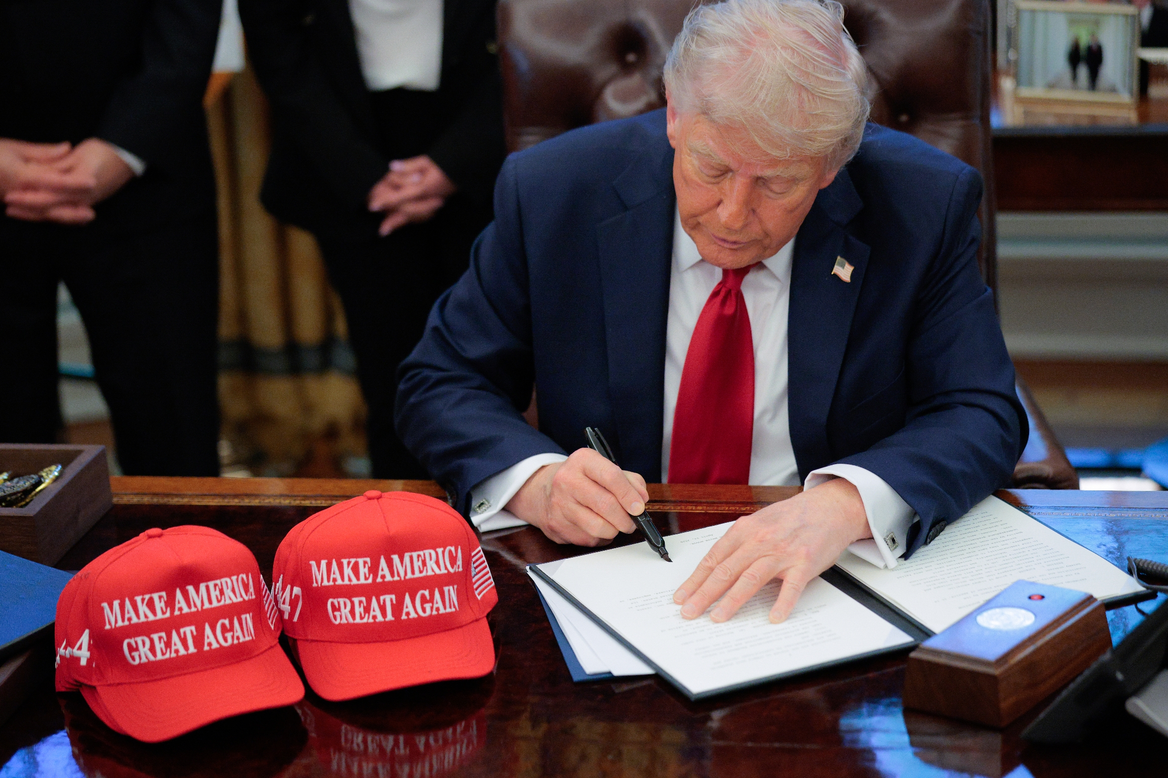 A man in a suit signs a document at a desk with two "Make America Great Again" hats and a large folder