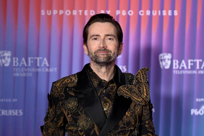A person poses on the BAFTA red carpet, wearing an ornate, patterned suit with gold detailing