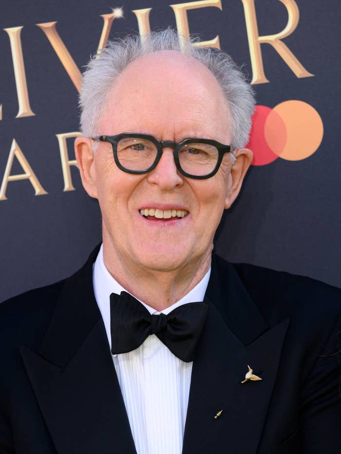 John Lithgow in glasses and tuxedo with bow tie standing in front of event backdrop, smiling