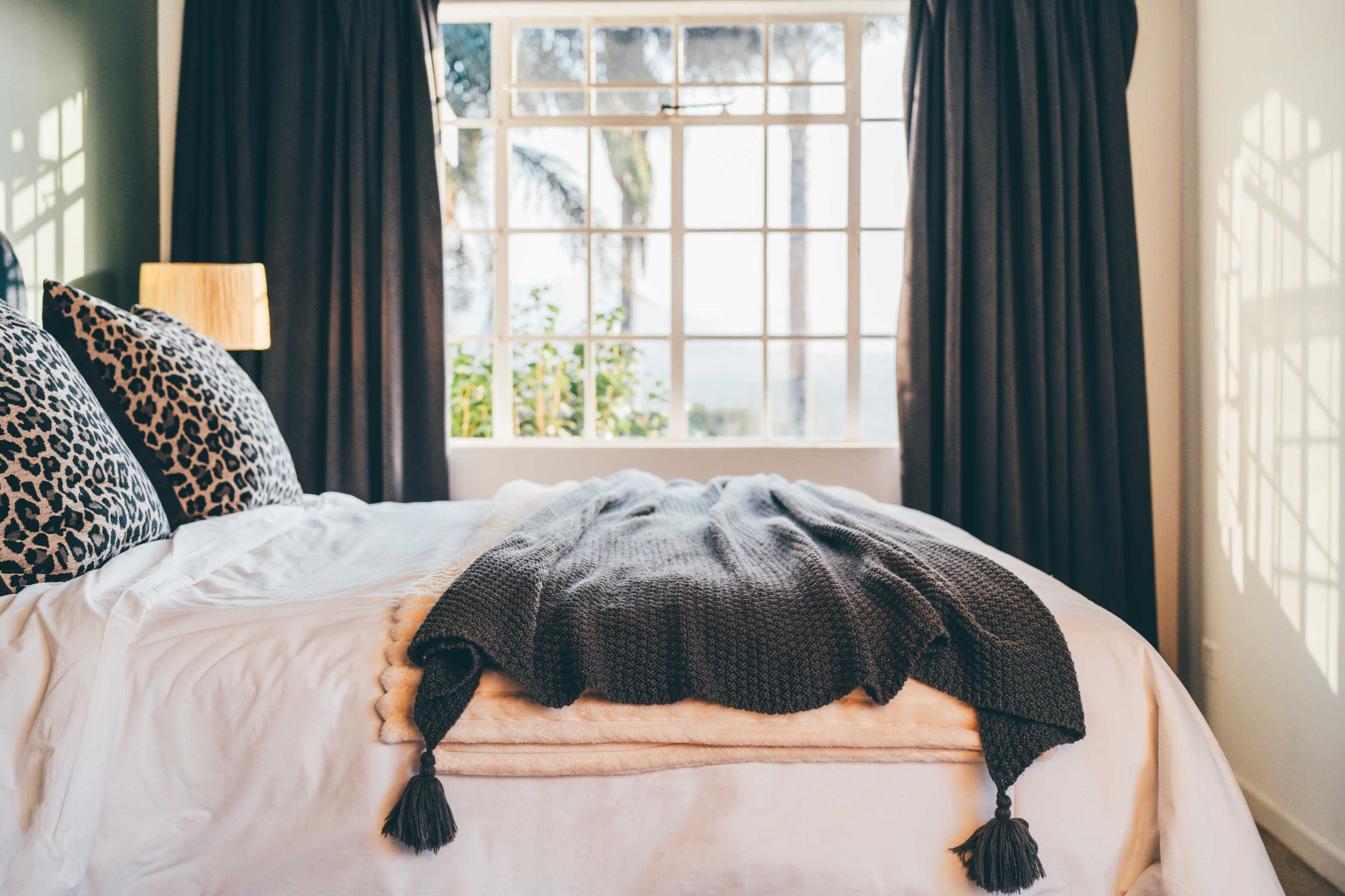 Cozy bedroom with patterned pillows and a throw on a neatly made bed, large window view with curtains framing trees and distant scenery.