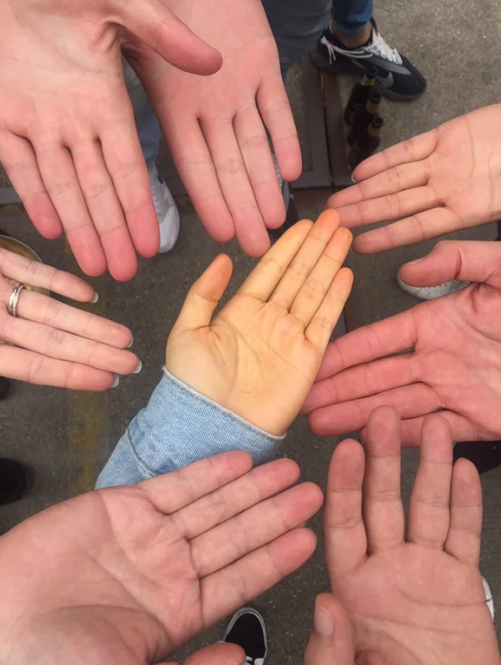Seven hands of various skin tones form a circle around a hand with an orange tint