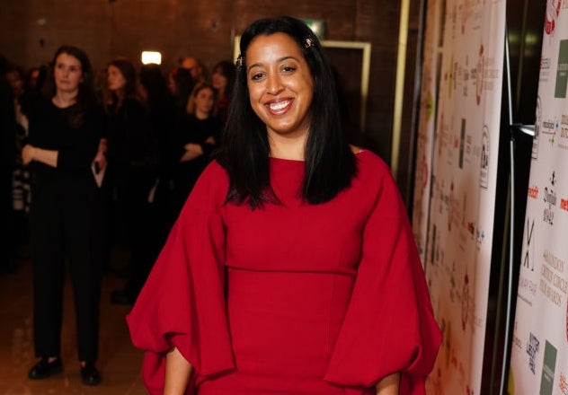 Person smiling on a red carpet, wearing a long red and white dress with flowing sleeves and shiny shoes