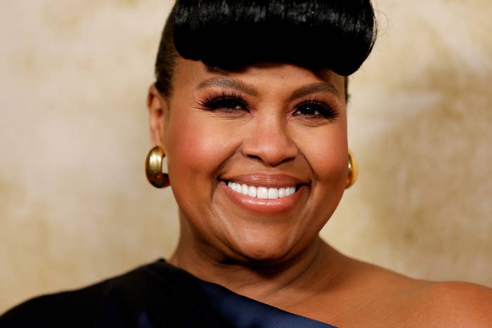 Person smiling, wearing a single-shoulder dress and gold hoop earrings. Hair styled with a voluminous front fringe