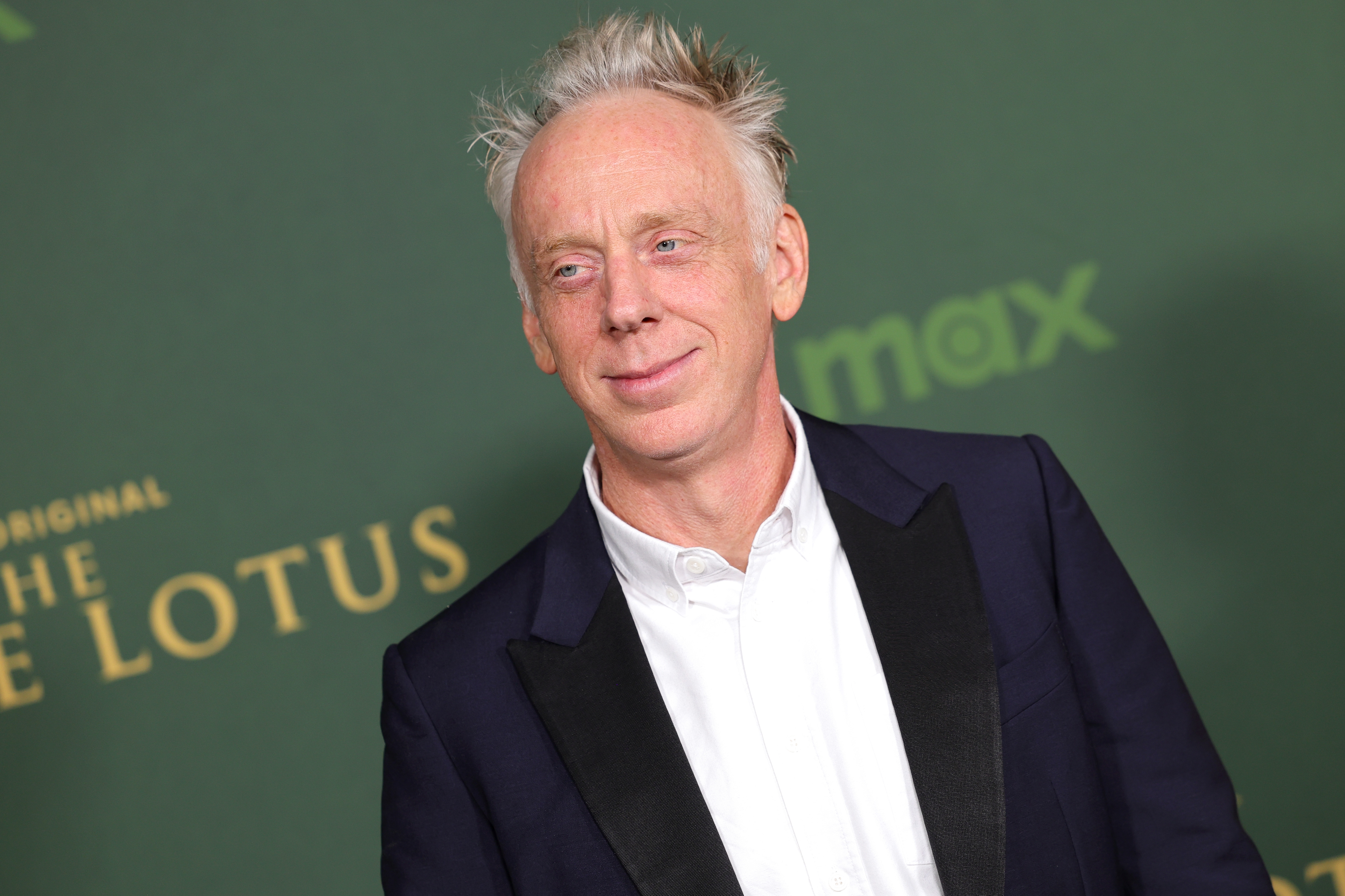 A person in a suit with a white shirt poses at an event backdrop with text "The White Lotus."