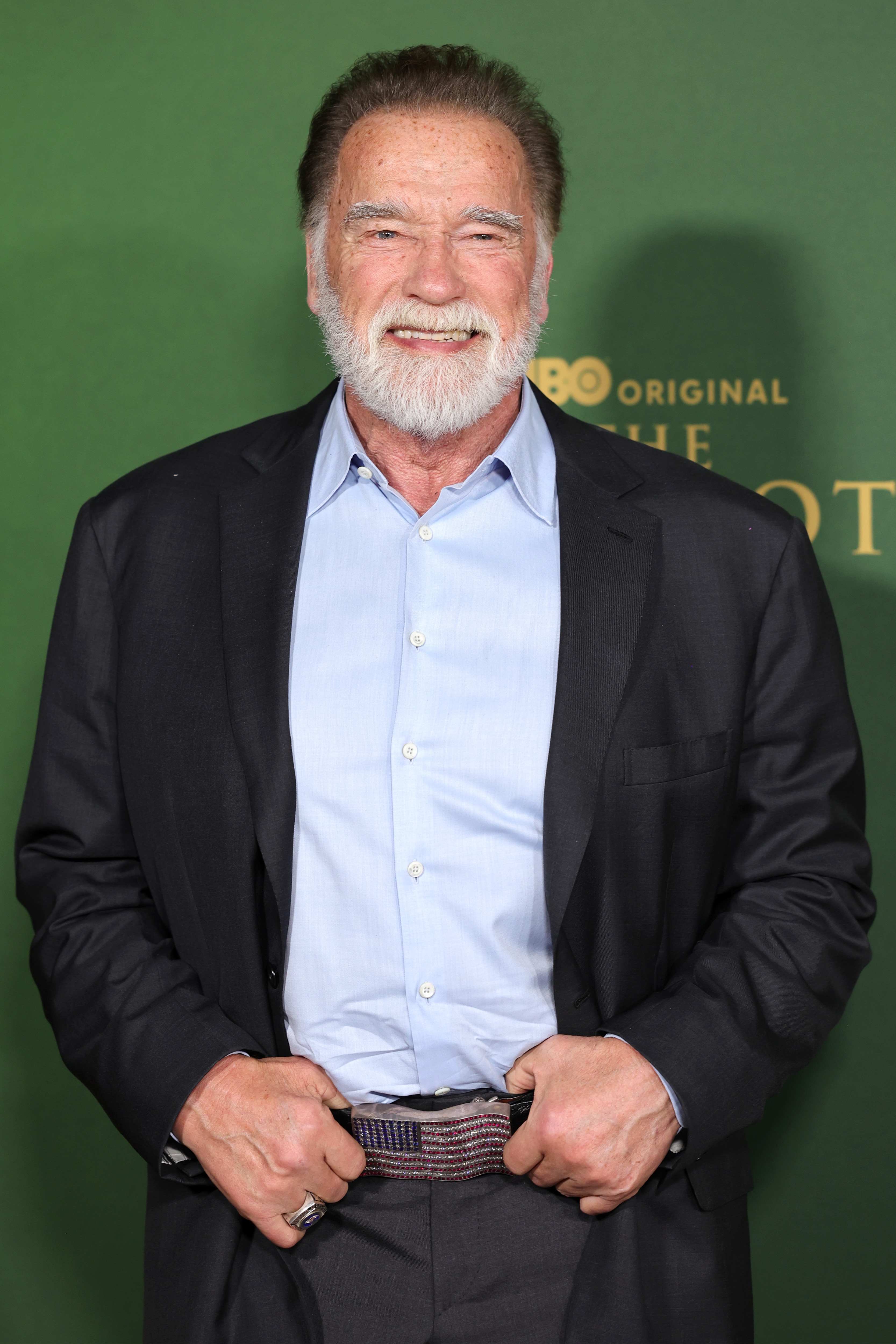 Man smiling at an event, wearing a dark blazer over a light shirt, with hands holding a belt