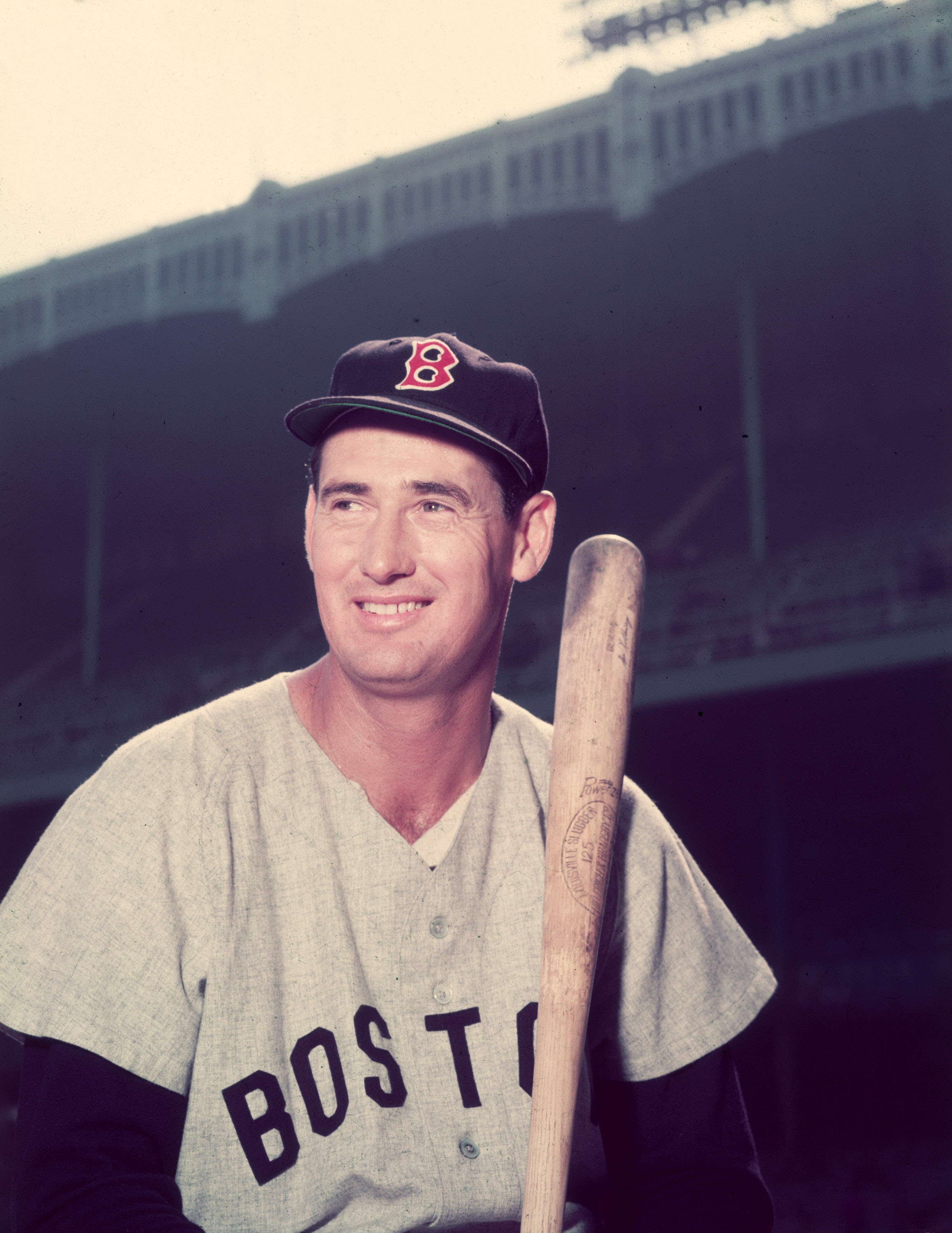A baseball player holding a bat, wearing a uniform with "Boston" on the jersey and a cap with a "B" emblem. Stadium seats are visible in the background