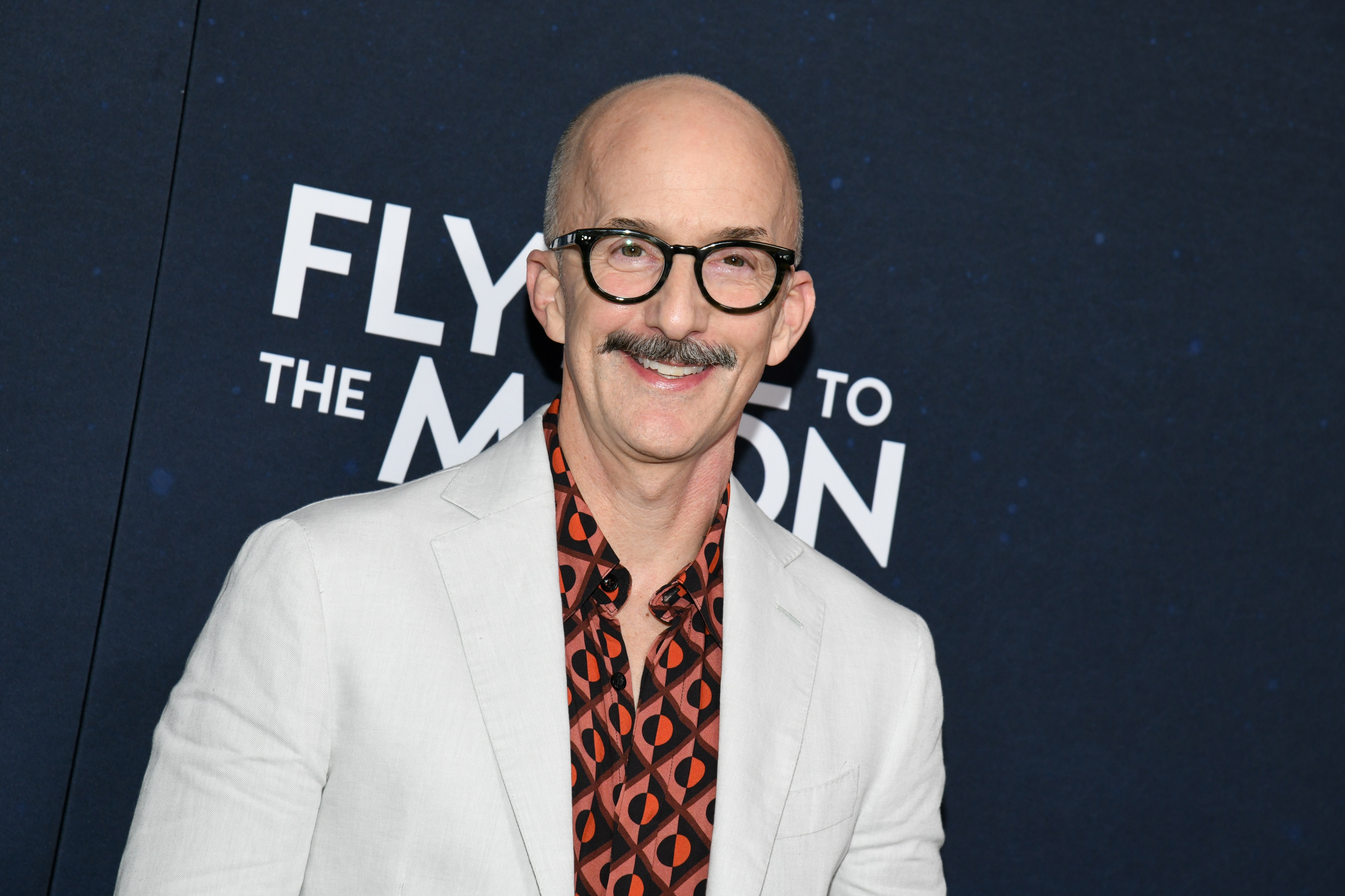 Person smiling at an event, wearing a patterned shirt under a light blazer, standing in front of a backdrop with the text "FLY TO THE MOON."