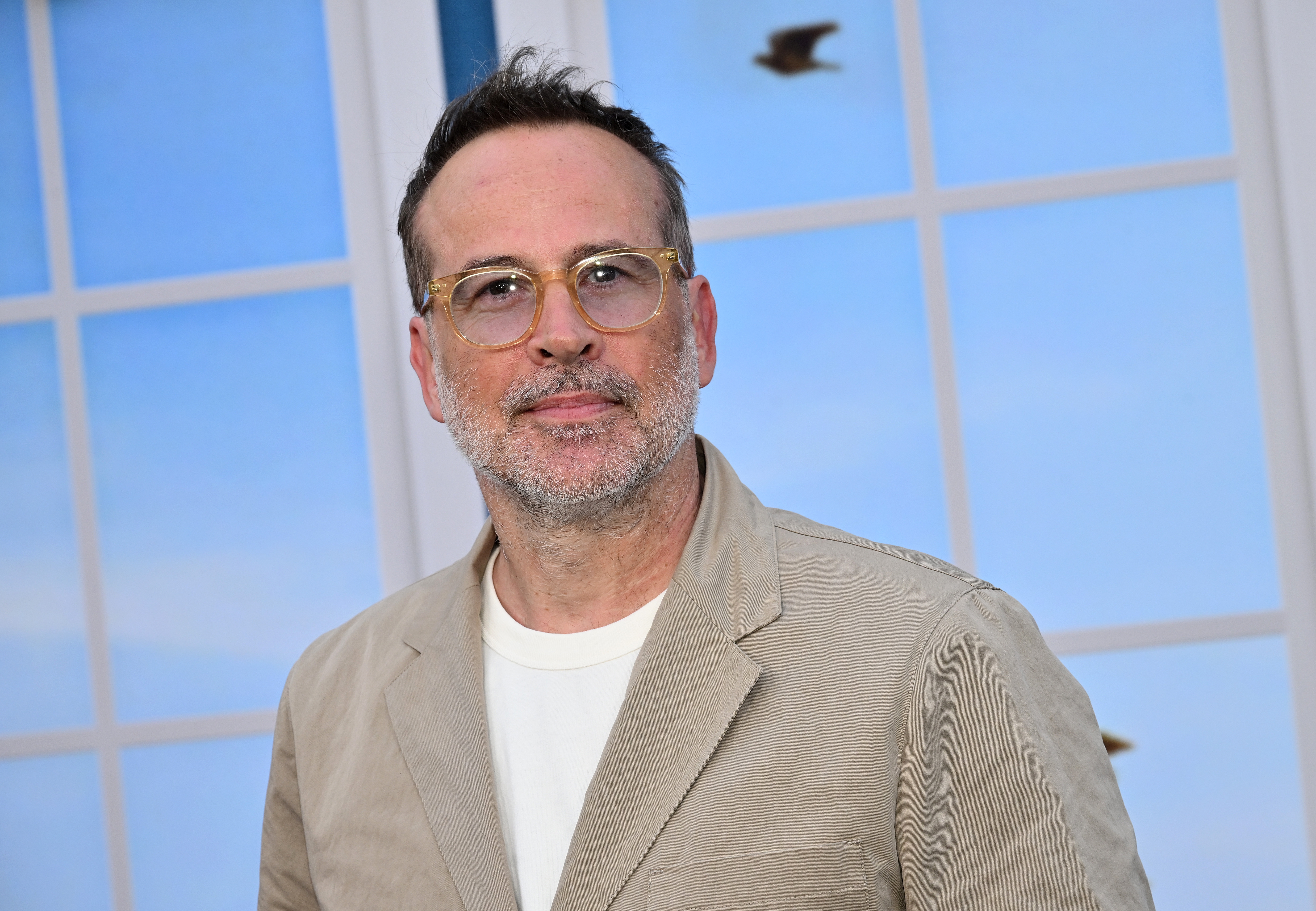 Person with short hair and glasses, wearing a light suit jacket over a white shirt, standing in front of a window with a blue sky